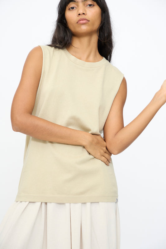 A woman wears the Extreme Cashmere No. 408 Muscle Tee in Desert with a light skirt, standing against a plain background, one hand on her waist and the other raised.