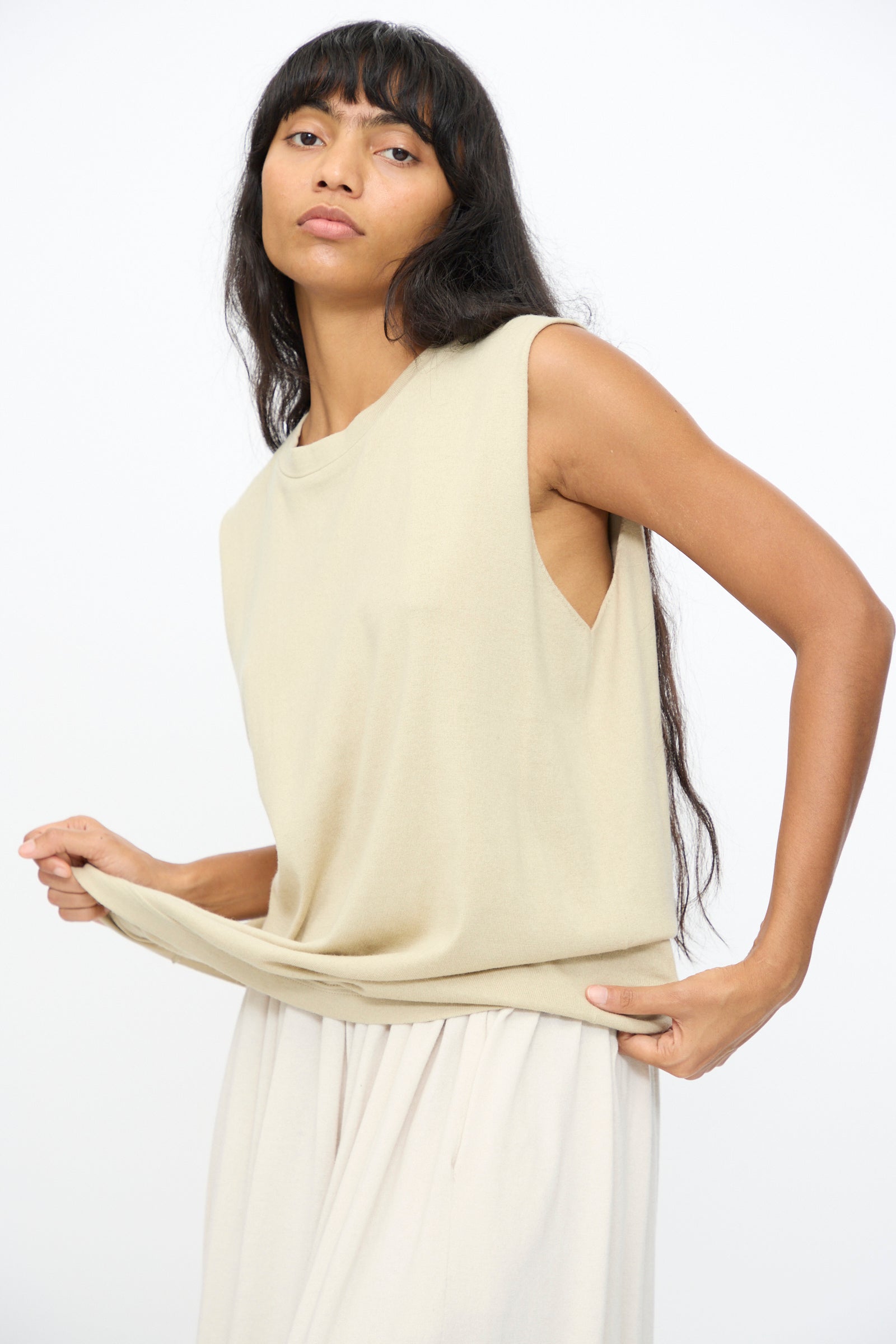A person with long dark hair wears the Extreme Cashmere No. 408 Muscle Tee in Desert, a sleeveless beige cotton-cashmere blend top, paired with a matching skirt and holds the hem of the top against a plain white background.