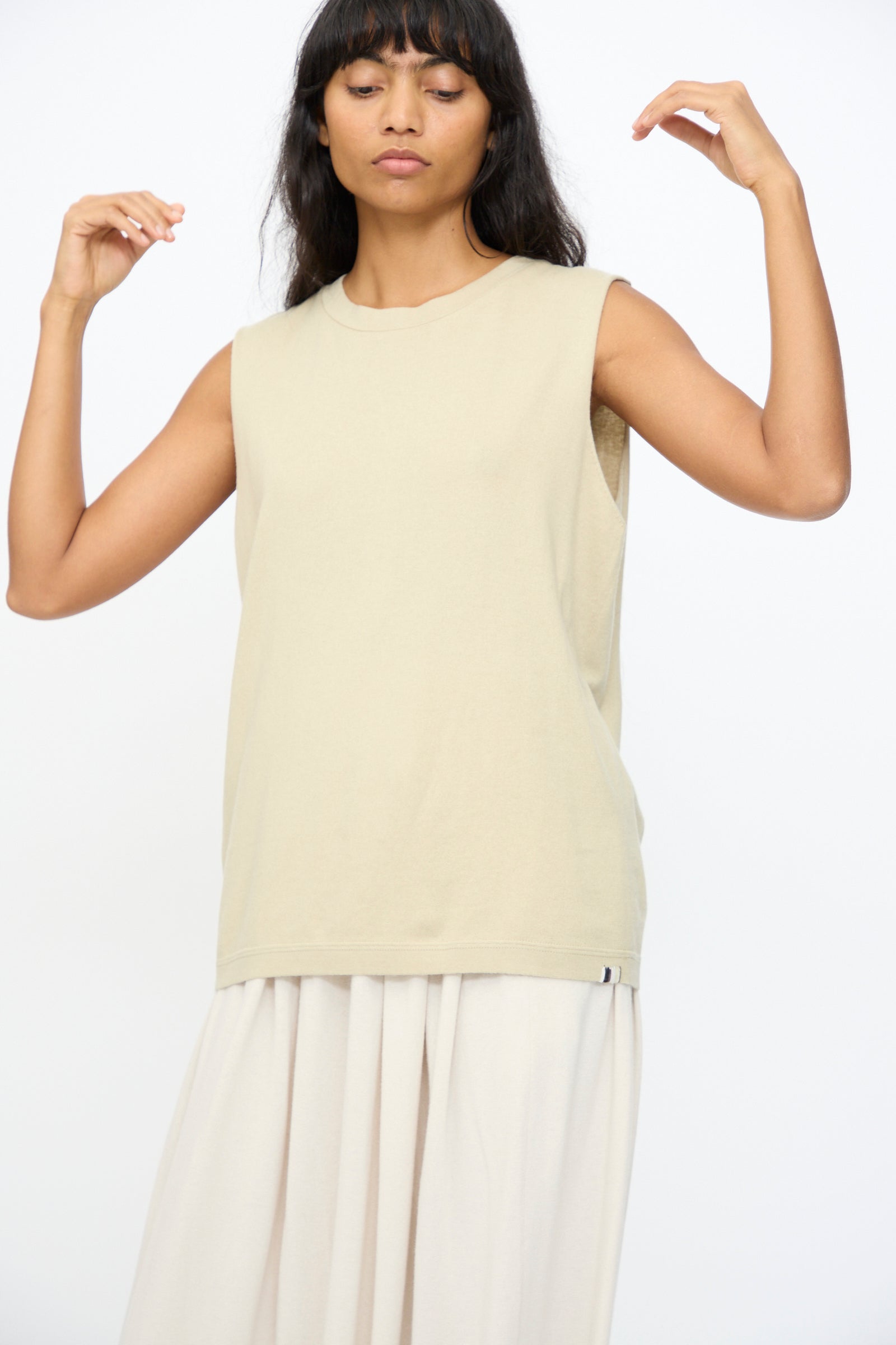 A person wears the Extreme Cashmere No. 408 Muscle Tee in Desert and a light pleated skirt, standing with arms slightly raised against a plain white background.