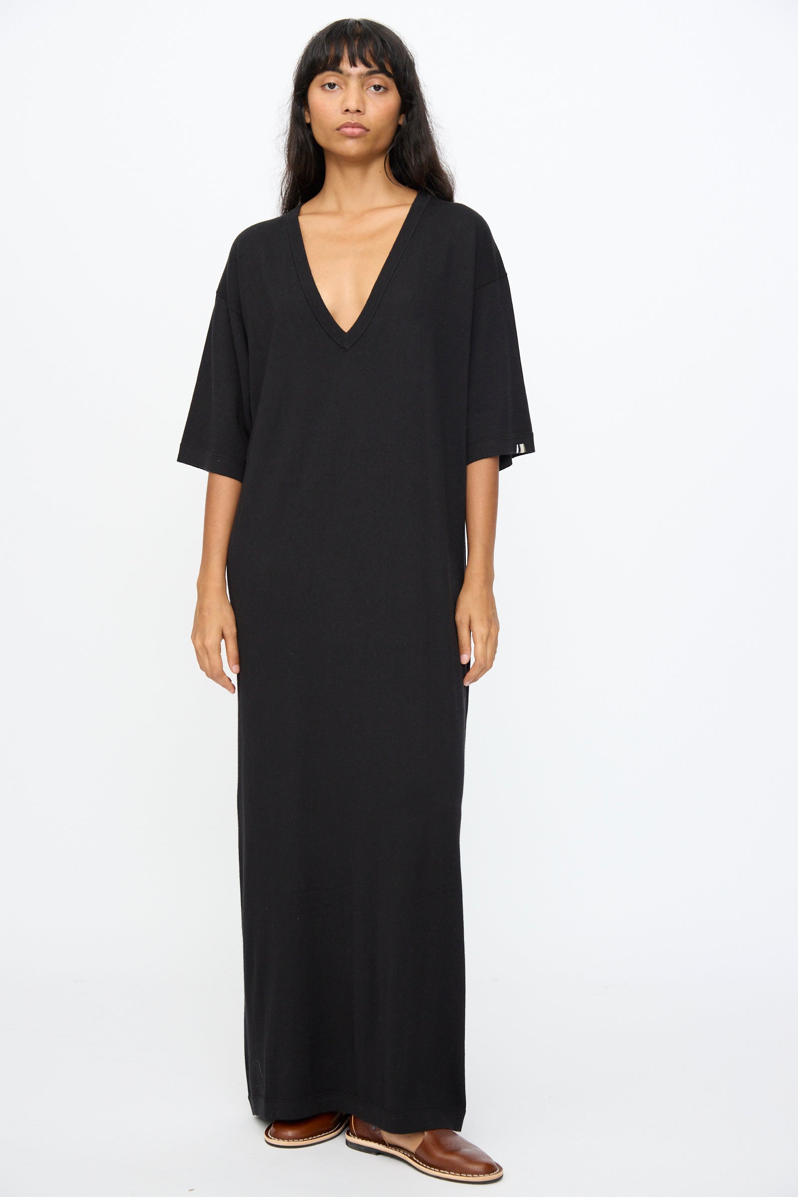 A person stands against a plain background wearing Extreme Cashmere's No. 410 Inda Dress in Raven—an ankle-length, relaxed-fit black t-shirt dress with short sleeves and a deep V-neck—paired with brown shoes.