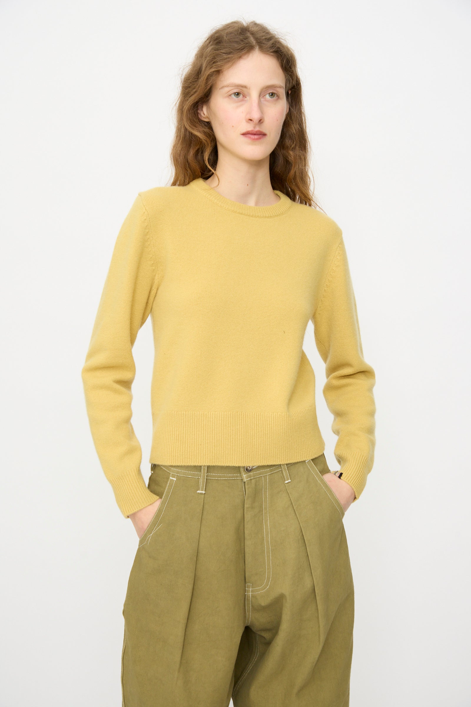A person with long wavy hair wears the Extreme Cashmere No. 430 Sweater in Corn and olive green pants, standing against a plain white background with hands in pockets.