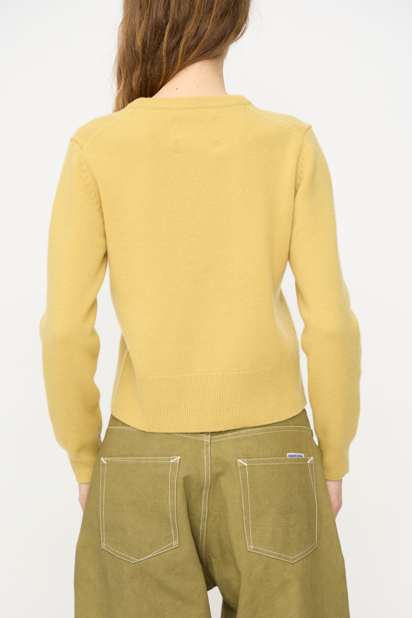 A person stands facing away, wearing the Extreme Cashmere No. 430 Sweater in Corn and olive green pants with visible white stitching on the back pockets.