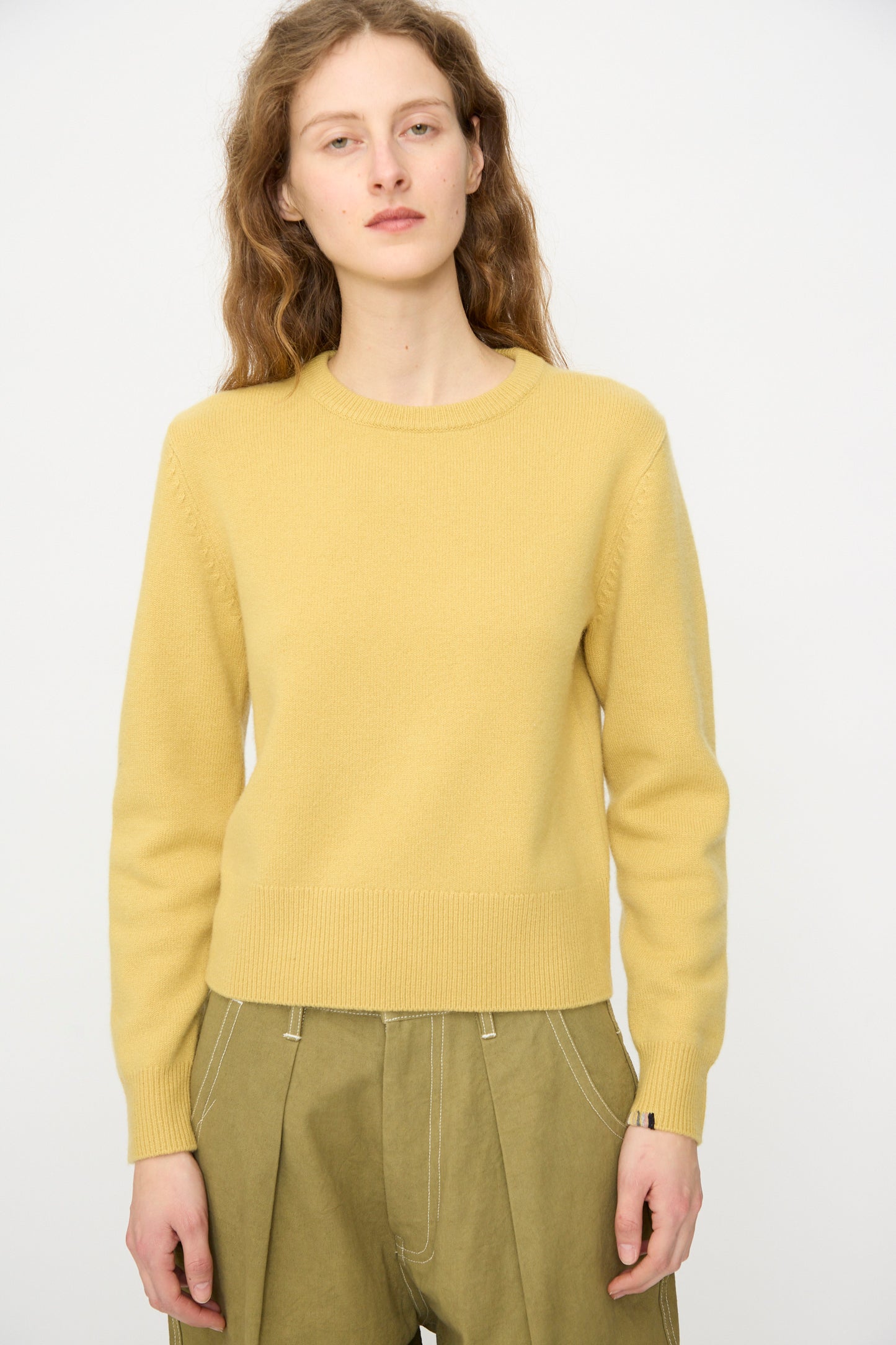 A person with long wavy hair wears the Extreme Cashmere No. 430 Sweater in Corn and olive green pants, standing against a plain white background.