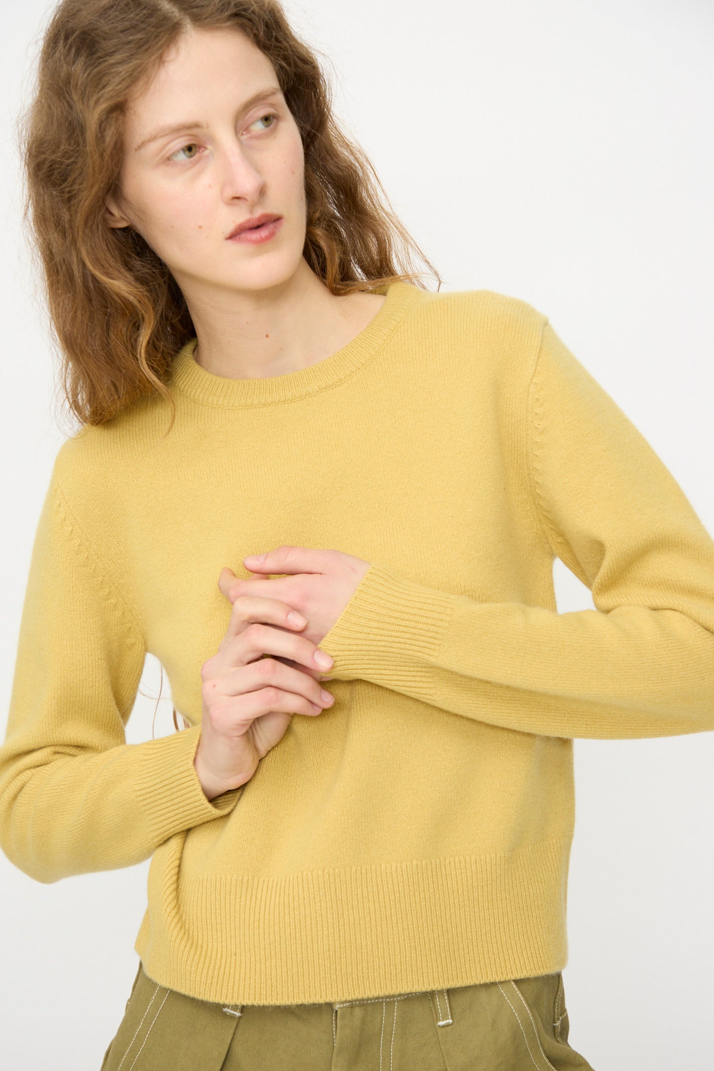 A person with long wavy hair wearing the Extreme Cashmere No. 430 Sweater in Corn and olive green pants stands against a plain white background.