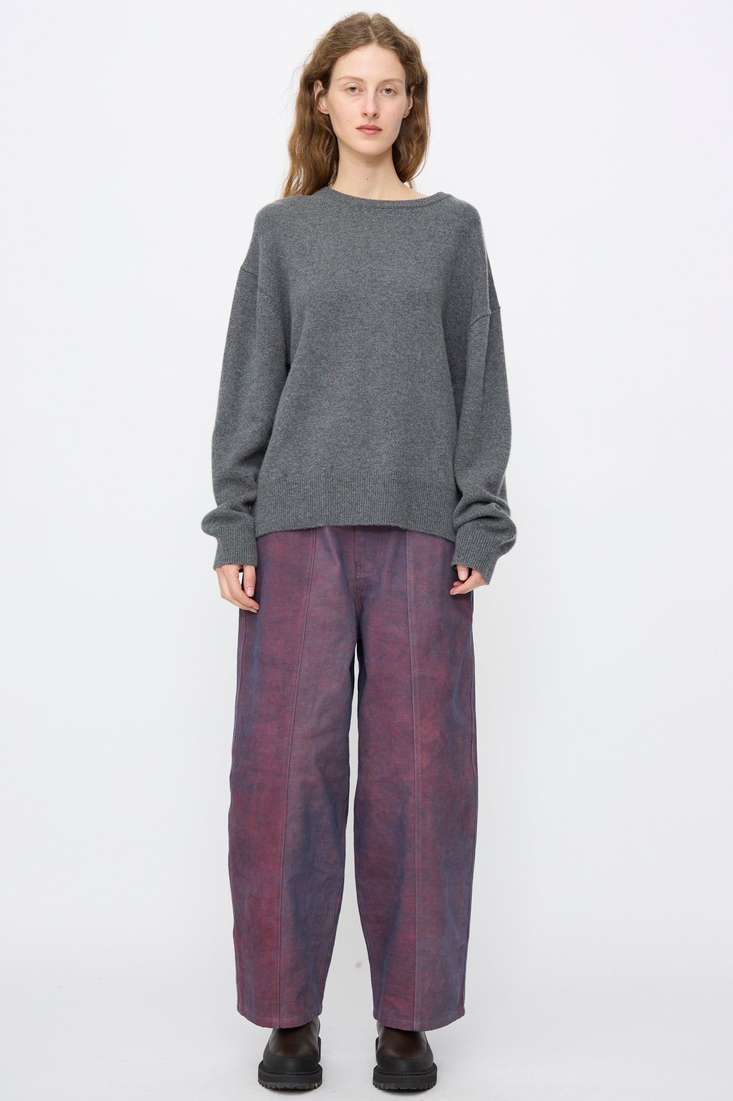 A person stands against a plain background wearing the Extreme Cashmere No. 431 Mike Sweater in Felt, wide-leg purple pants, and black shoes.