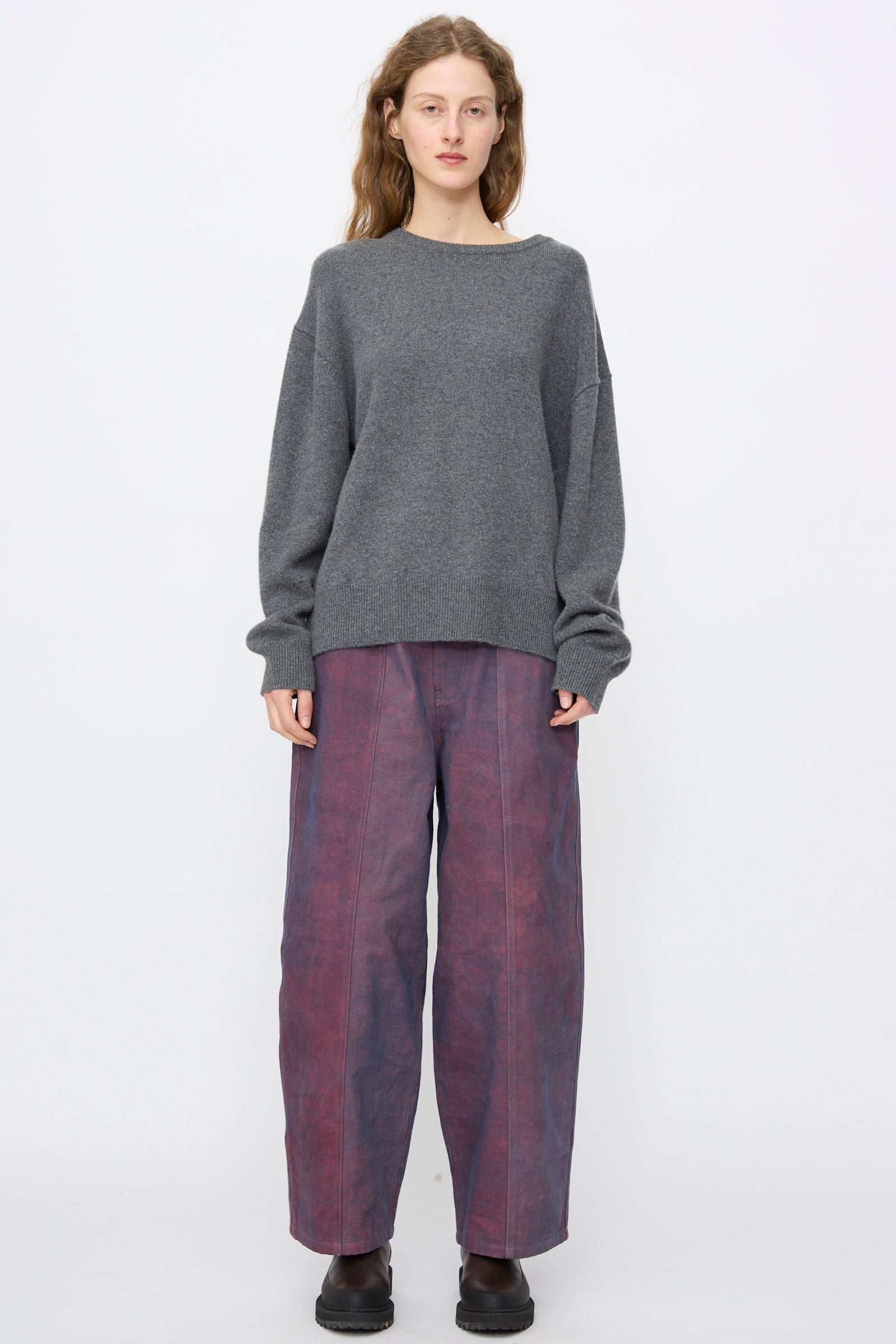 A person stands against a plain background wearing the Extreme Cashmere No. 431 Mike Sweater in Felt, wide-leg purple pants, and black shoes.