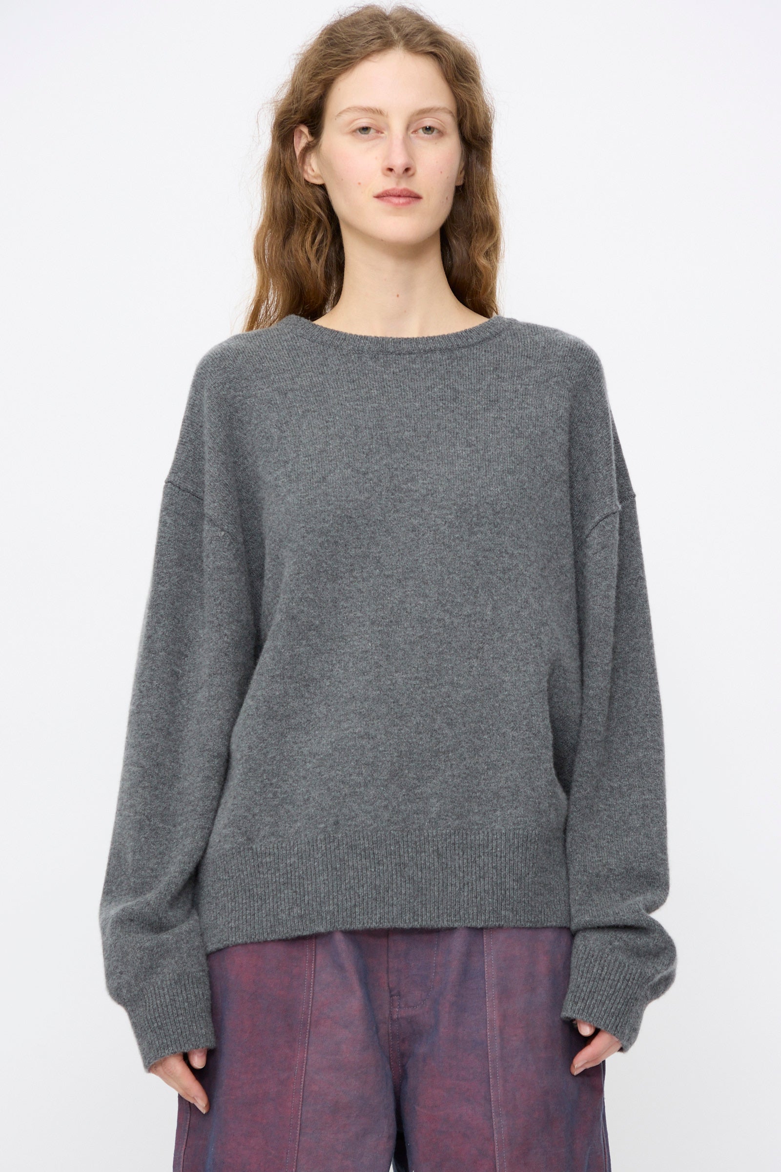 A person with long, wavy hair wears the Extreme Cashmere No. 431 Mike Sweater in Felt (grey), paired with purple pants, standing against a plain white background.
