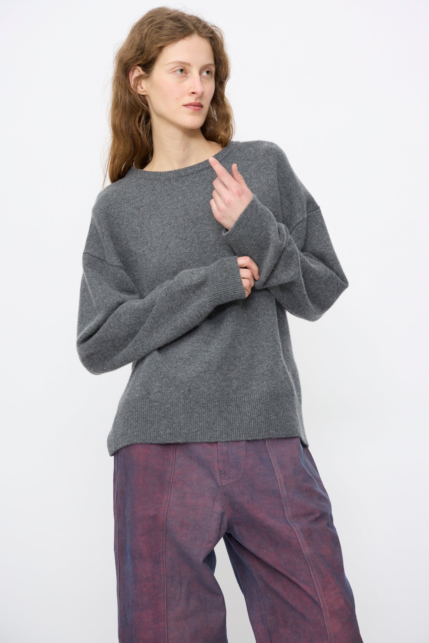 A person with long hair wears the Extreme Cashmere No. 431 Mike Sweater in Felt, a relaxed-fit grey crewneck, paired with loose purple pants, standing against a plain white background.