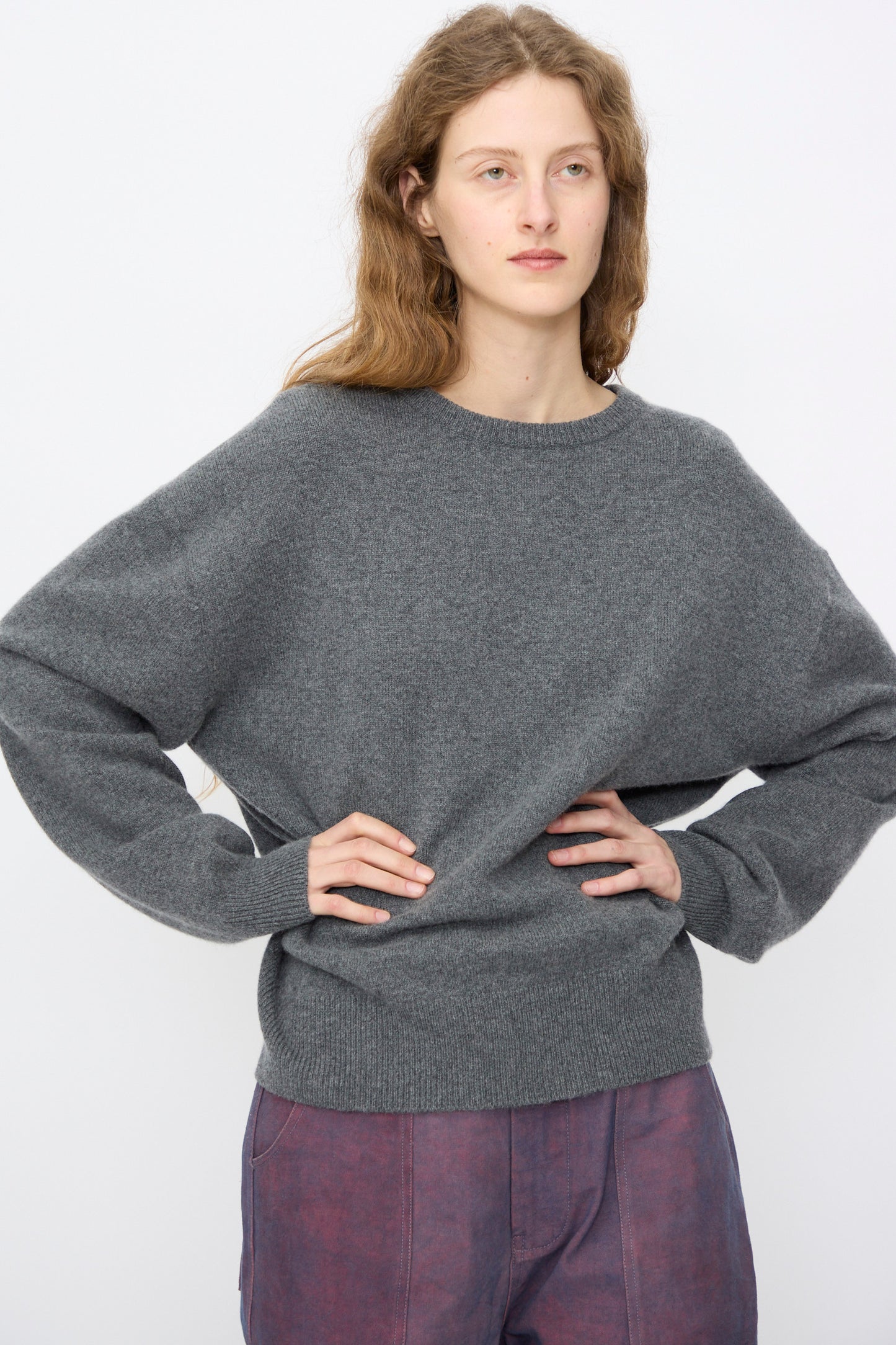 A person with long, wavy hair wears the Extreme Cashmere No. 431 Mike Sweater in Felt, paired with purple pants, standing against a plain light background with hands on hips.