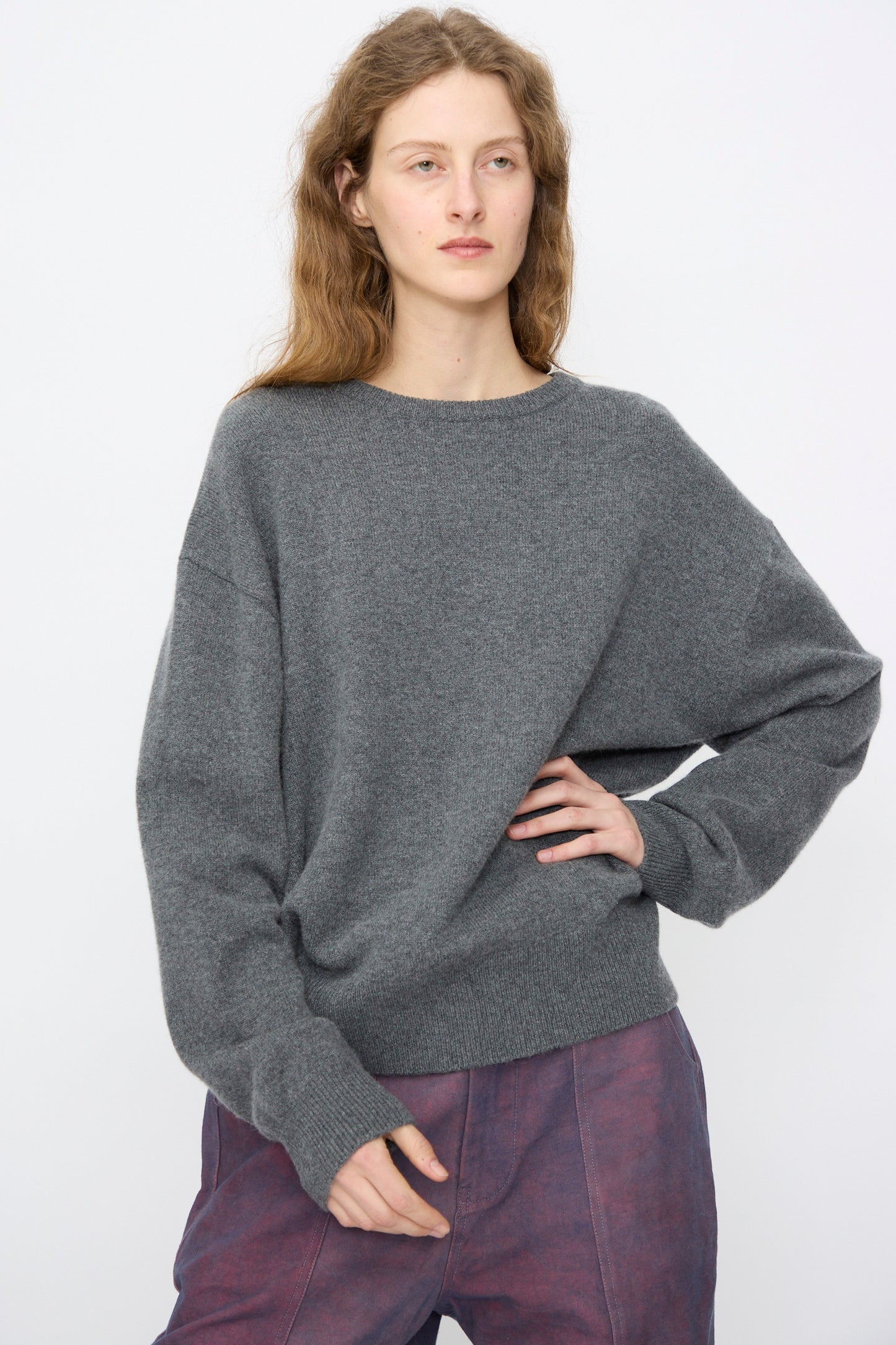 A person with long wavy hair wears the Extreme Cashmere No. 431 Mike Sweater in Felt (gray) and purple pants, standing against a plain light background.