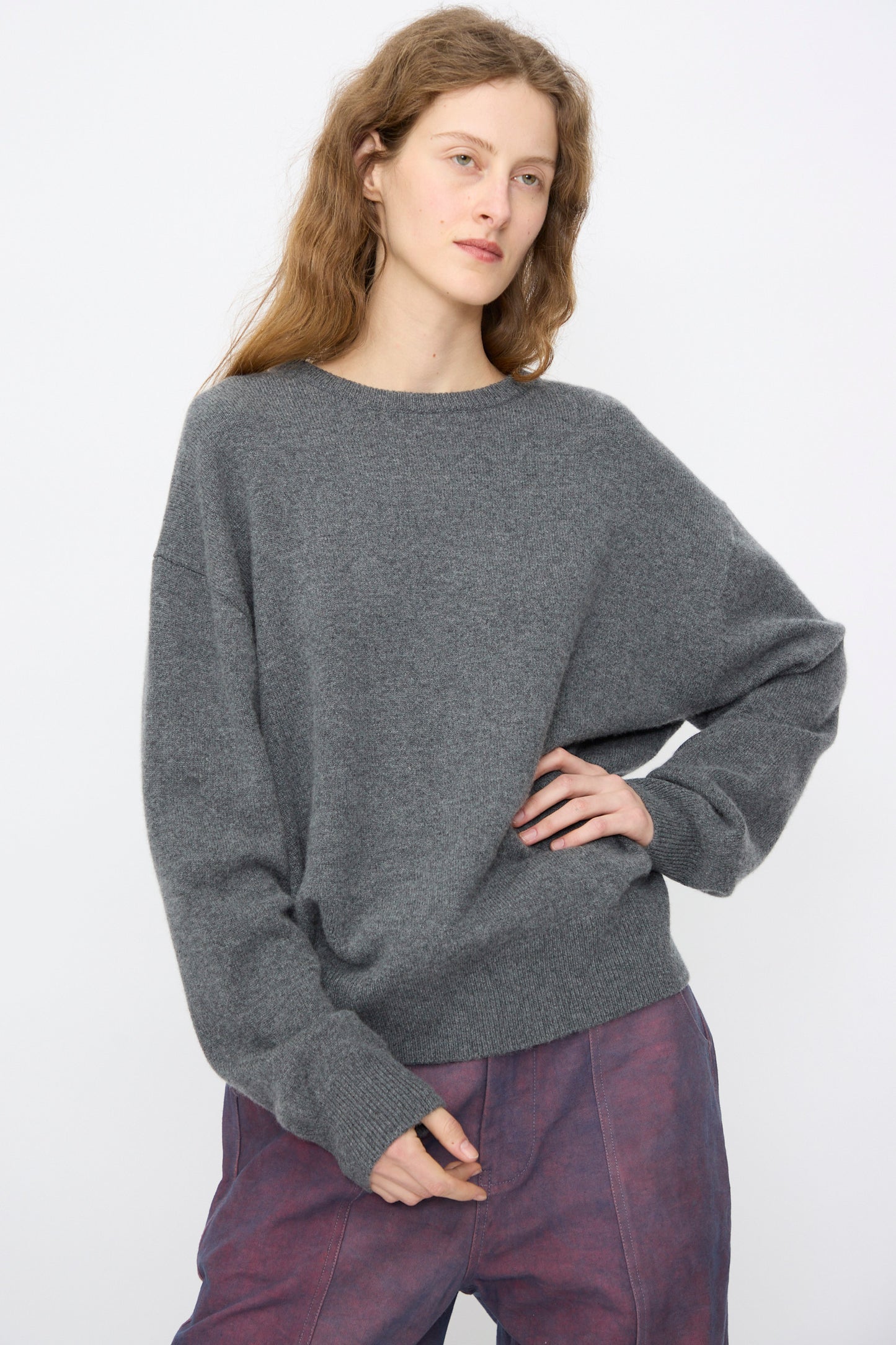 A woman with long, light brown hair stands against a plain background, wearing Extreme Cashmere's No. 431 Mike Sweater in Felt—a relaxed-fit, machine washable gray crewneck—paired with purple pants for effortless comfort and ease.