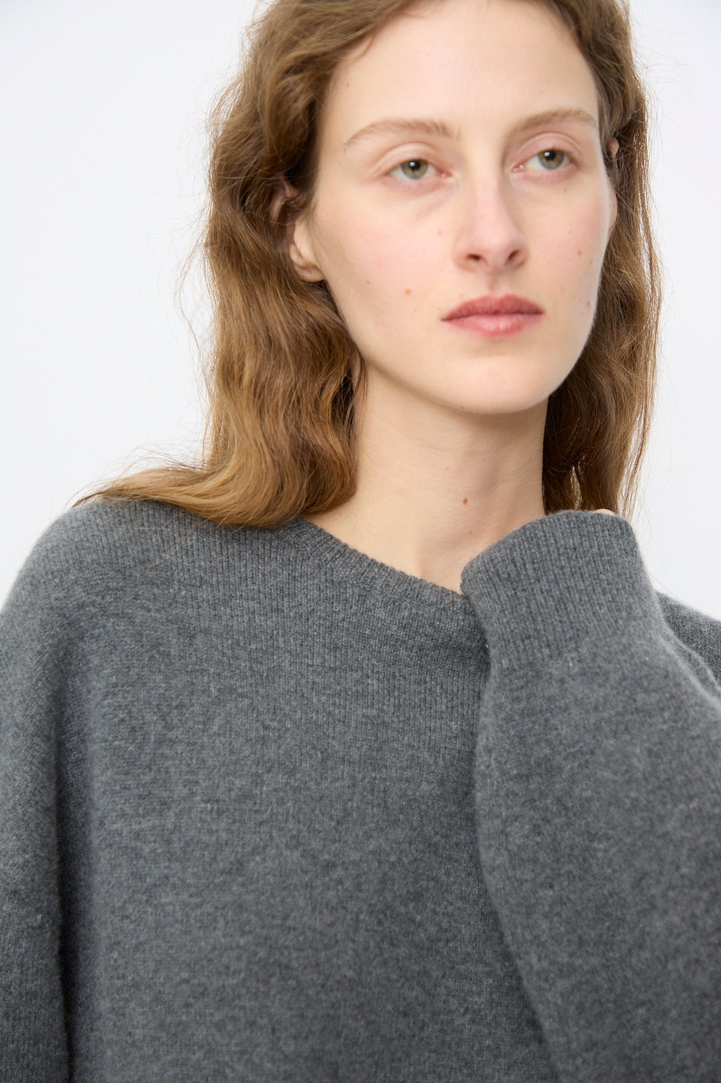 A person with long, wavy light brown hair models the No. 431 Mike Sweater in Felt by Extreme Cashmere—a relaxed-fit gray crewneck—while gazing to the side against a simple background.
