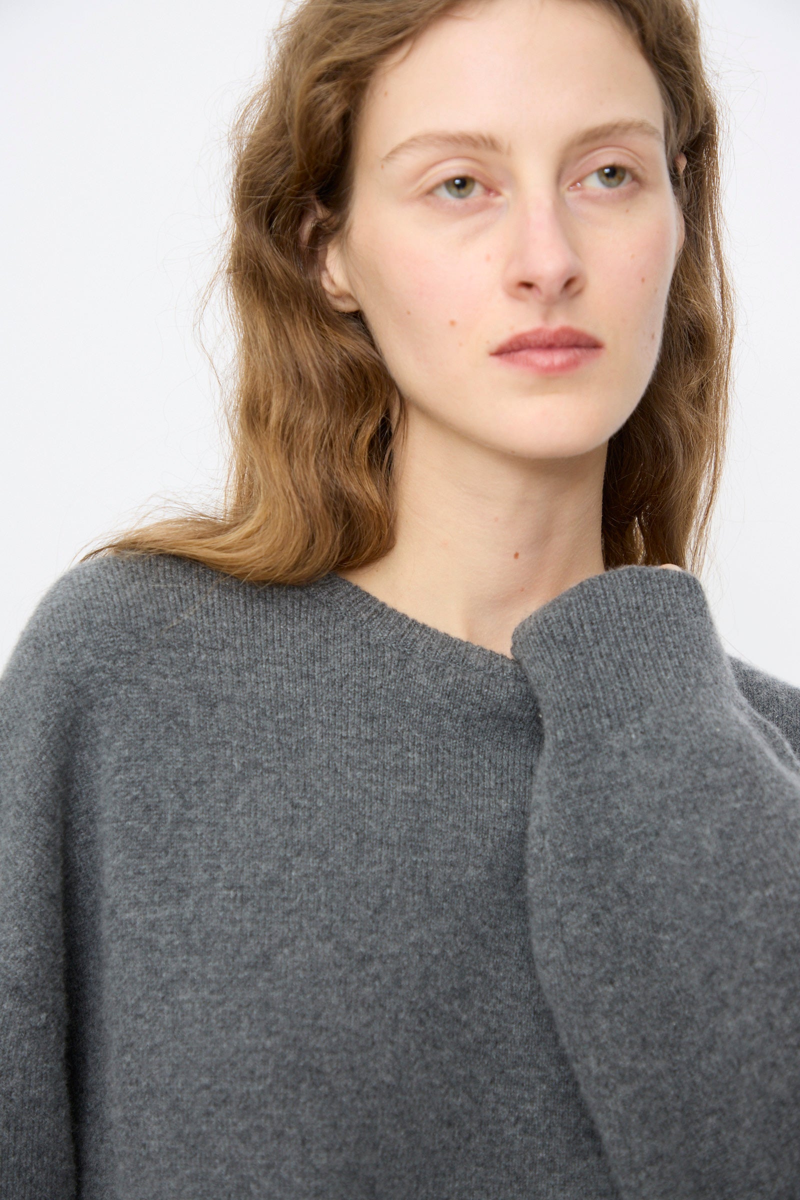 A person with long, wavy light brown hair models the No. 431 Mike Sweater in Felt by Extreme Cashmere—a relaxed-fit gray crewneck—while gazing to the side against a simple background.