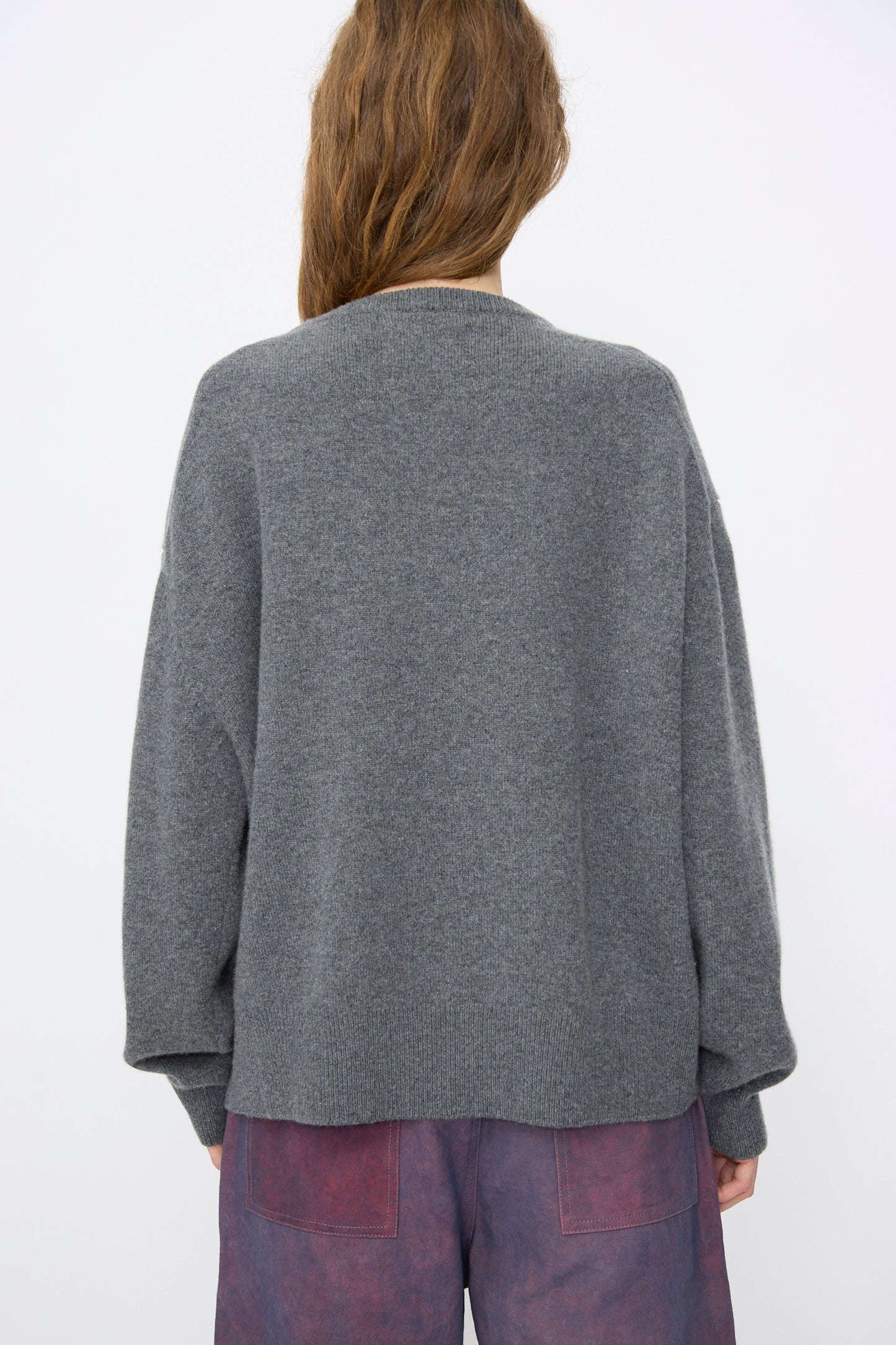 A person with long brown hair is shown from the back, wearing the Extreme Cashmere No. 431 Mike Sweater in Felt paired with purple pants, set against a plain light background.