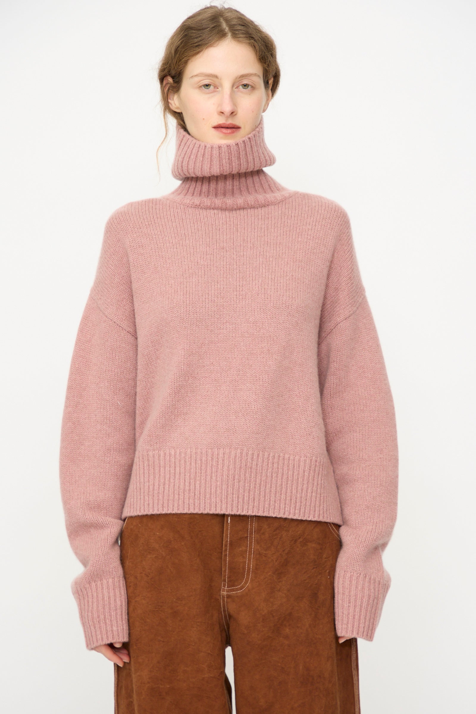 A person wears the Extreme Cashmere No. 434 Crop Xtra Sweater in Moor, a chunky pink cashmere turtleneck with long sleeves, paired with brown wide-legged pants, standing against a plain white background.
