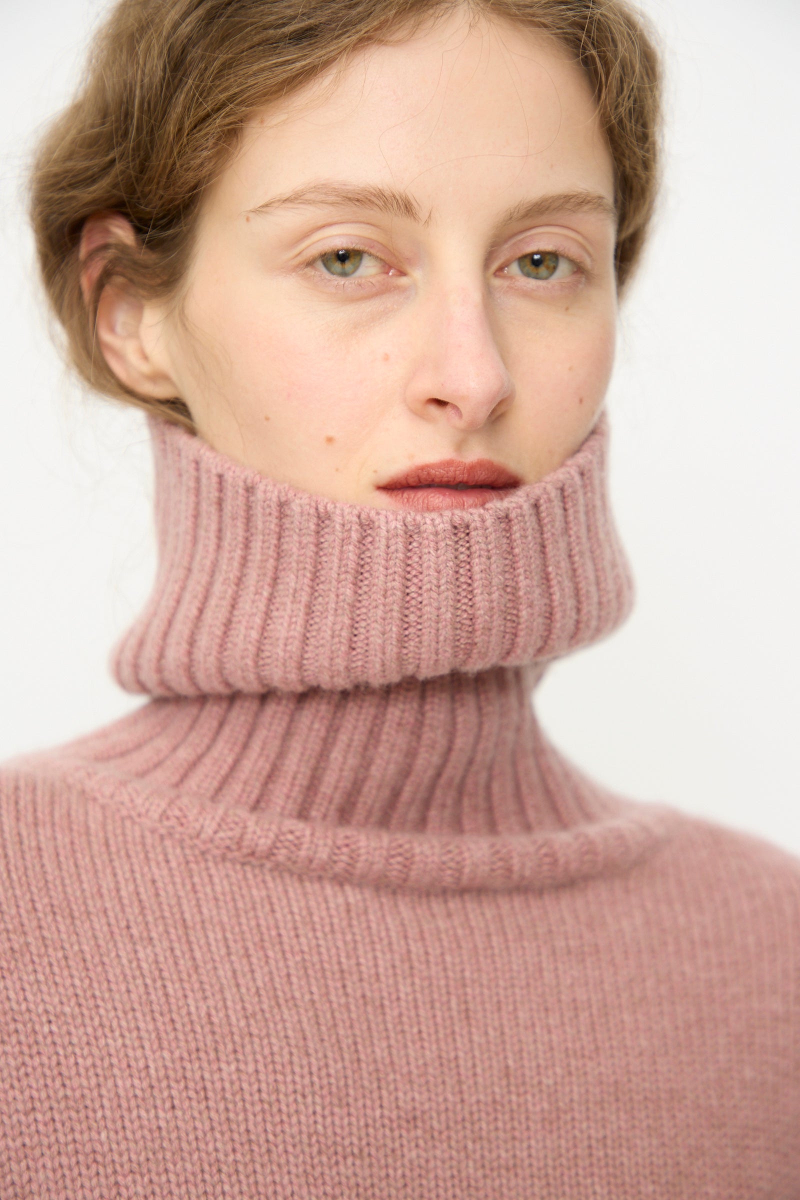 Person wearing the Extreme Cashmere No. 434 Crop Xtra Sweater in Moor, collar pulled up to cover the lower half of their face, looking at the camera.