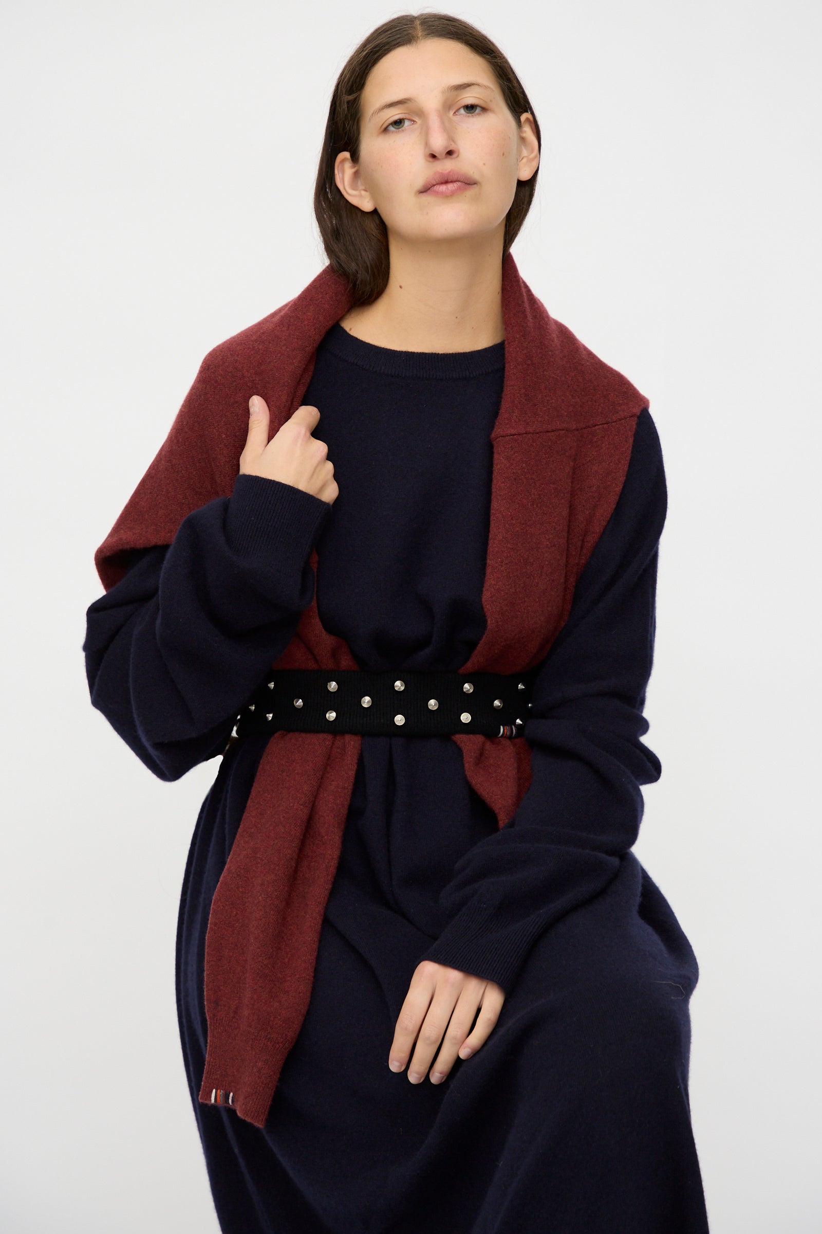 A person with straight brown hair sits against a plain light background, wearing a navy blue dress, a maroon scarf over their shoulders, and Extreme Cashmere's No. 446 Stud Belt.