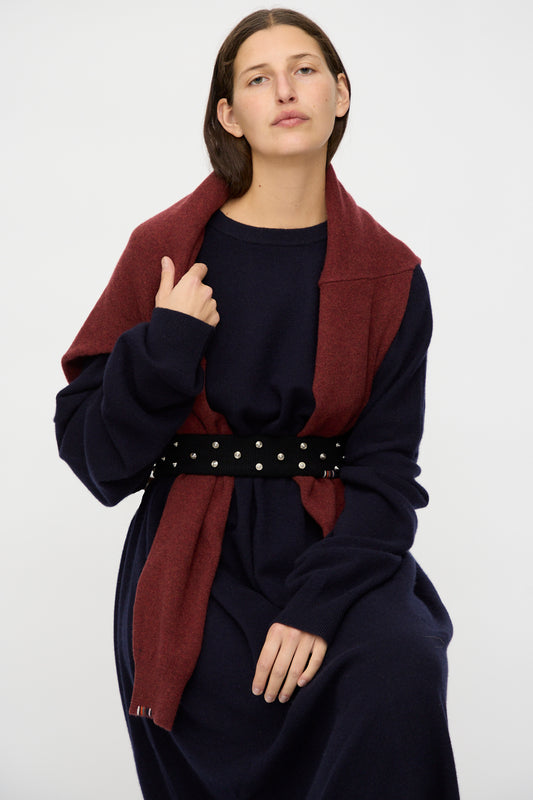 A person with straight brown hair sits against a plain light background, wearing a navy blue dress, a maroon scarf over their shoulders, and Extreme Cashmere's No. 446 Stud Belt.
