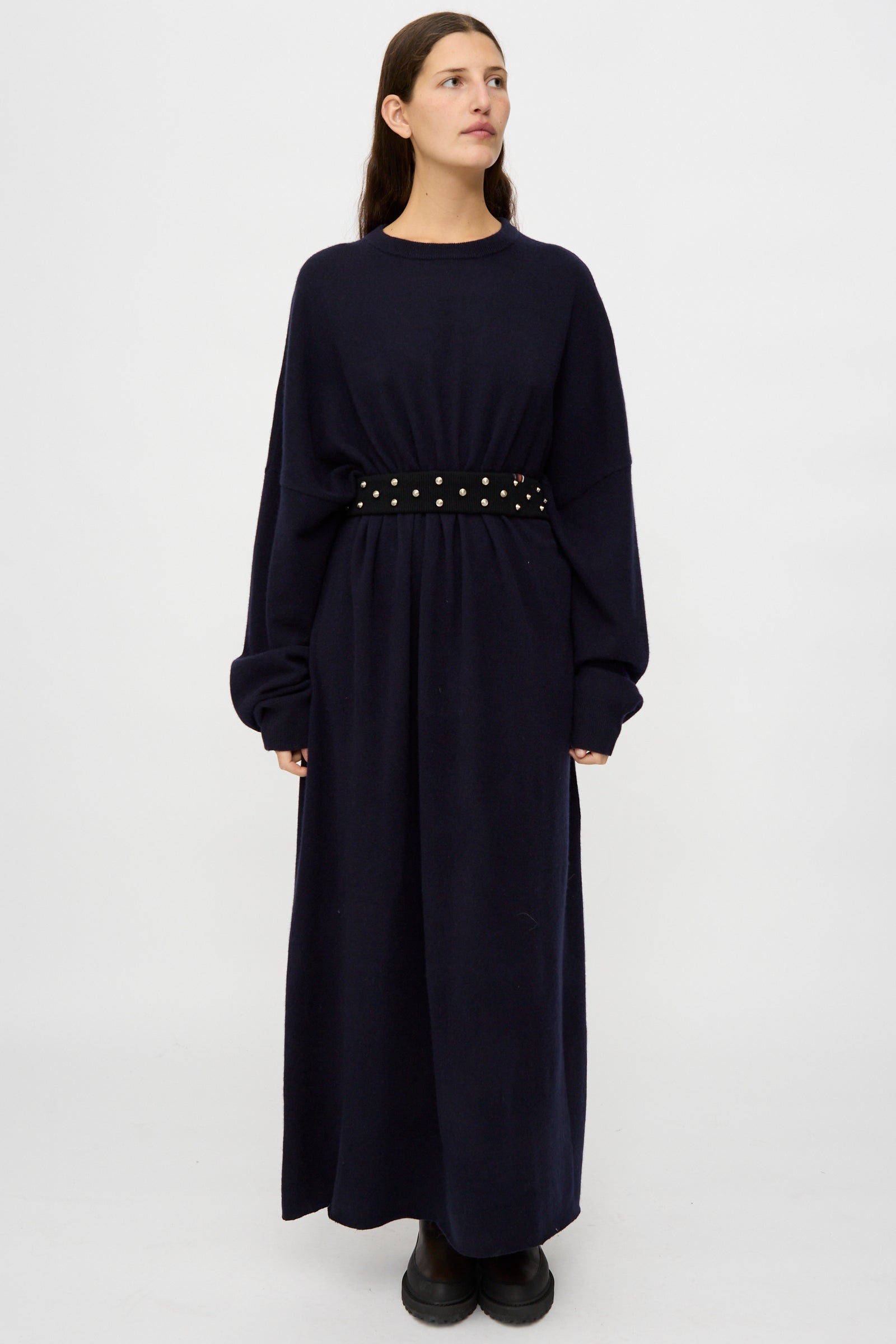 A person wearing a long, loose-fitting navy dress with the No. 446 Stud Belt by Extreme Cashmere and black boots stands against a plain white background.