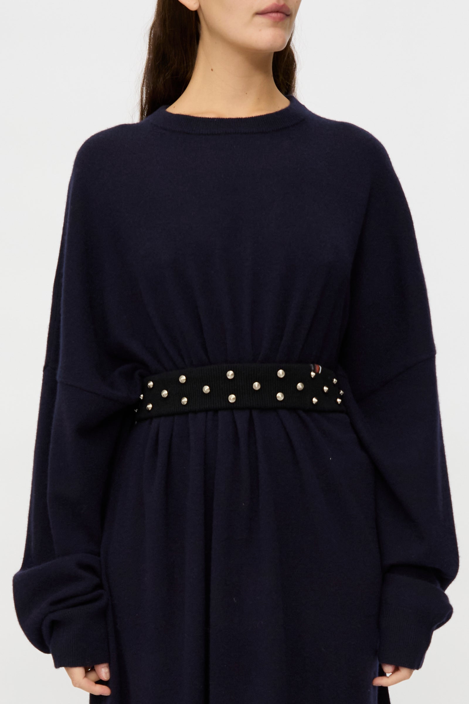 A person wears a loose-fitting, long-sleeved navy dress cinched at the waist with the Extreme Cashmere No. 446 Stud Belt for a bold accent.