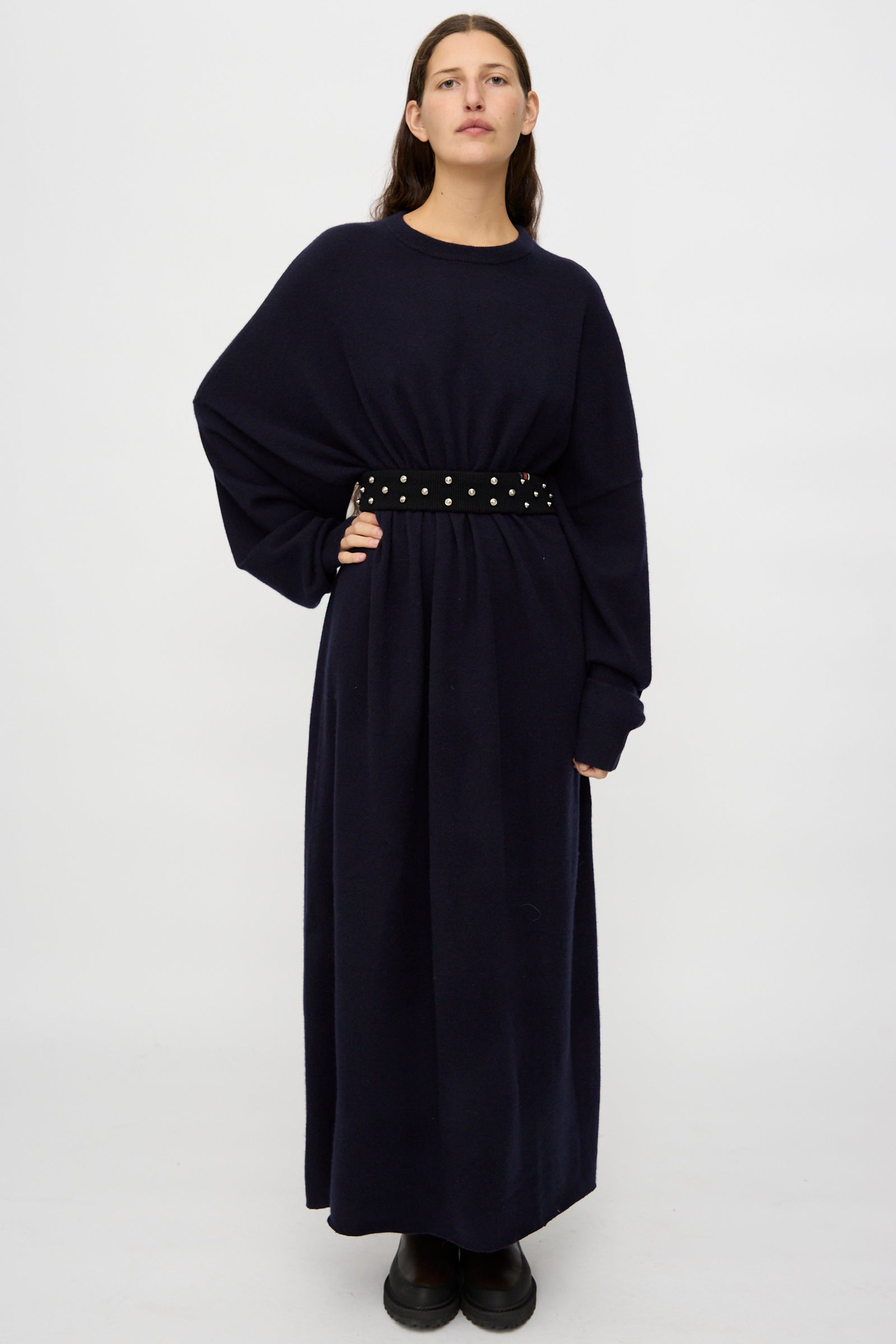 A person stands against a plain background wearing a long, dark oversized dress, cinched at the waist with the Extreme Cashmere No. 446 Stud Belt and paired with black shoes.