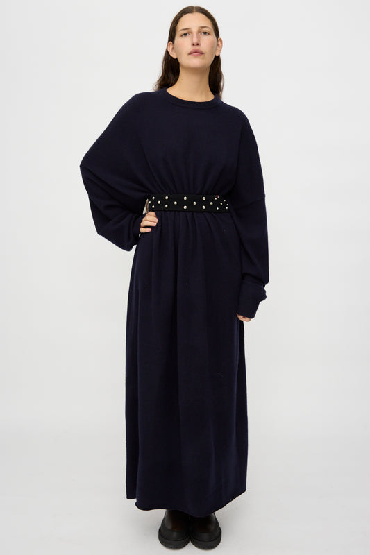 A person stands against a plain background wearing a long, dark oversized dress, cinched at the waist with the Extreme Cashmere No. 446 Stud Belt and paired with black shoes.