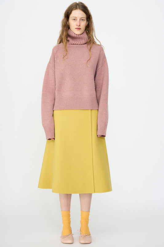 A woman stands against a plain white background wearing a pink turtleneck, the Extreme Cashmere No. 465 Line Long Skirt in Corn, orange socks, and beige shoes.