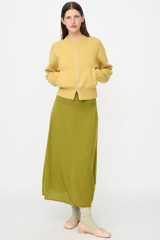 A person stands facing forward wearing a pale yellow zip-up jacket, the Extreme Cashmere No. 470 Seduce Skirt in Grass, light green socks, and pale pink shoes against a plain white background.