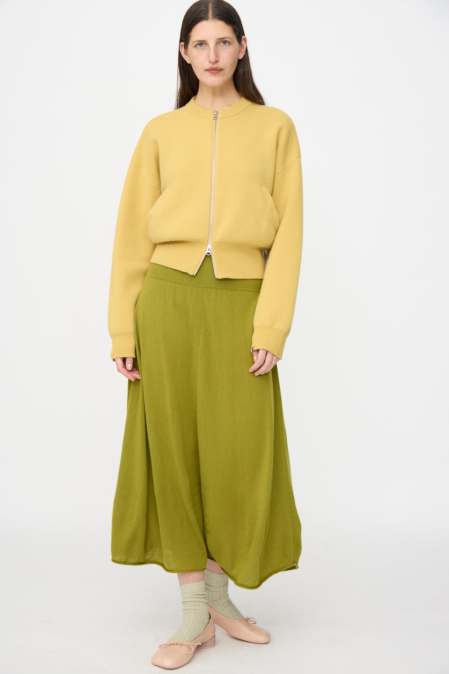 A woman stands against a plain background wearing a yellow zip-up jacket, the Extreme Cashmere No. 470 Seduce Skirt in Grass, and beige flats paired with light green socks.