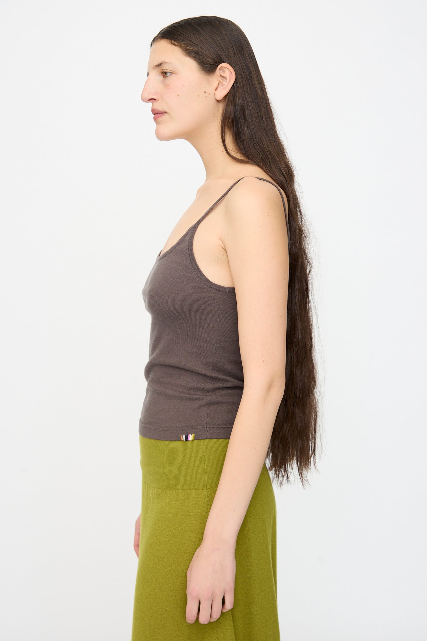 A person with long brown hair stands in profile wearing the Extreme Cashmere No. 471 Britney Tank in Mud, paired with a green skirt, against a plain white background.