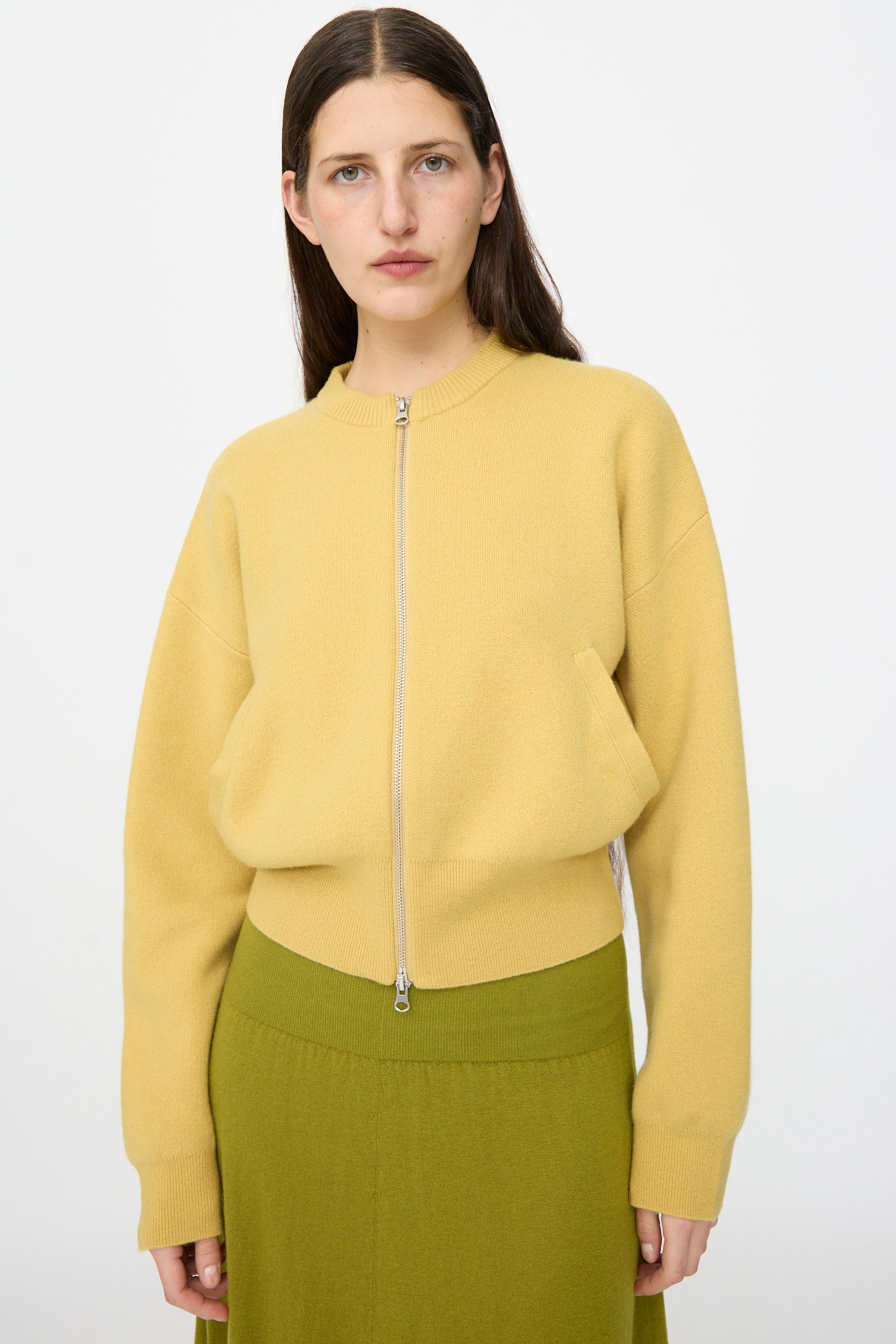 A woman with straight brown hair wears the Extreme Cashmere No. 478 Xtra Fun Cardigan in Corn and an olive green skirt, standing against a plain white background.