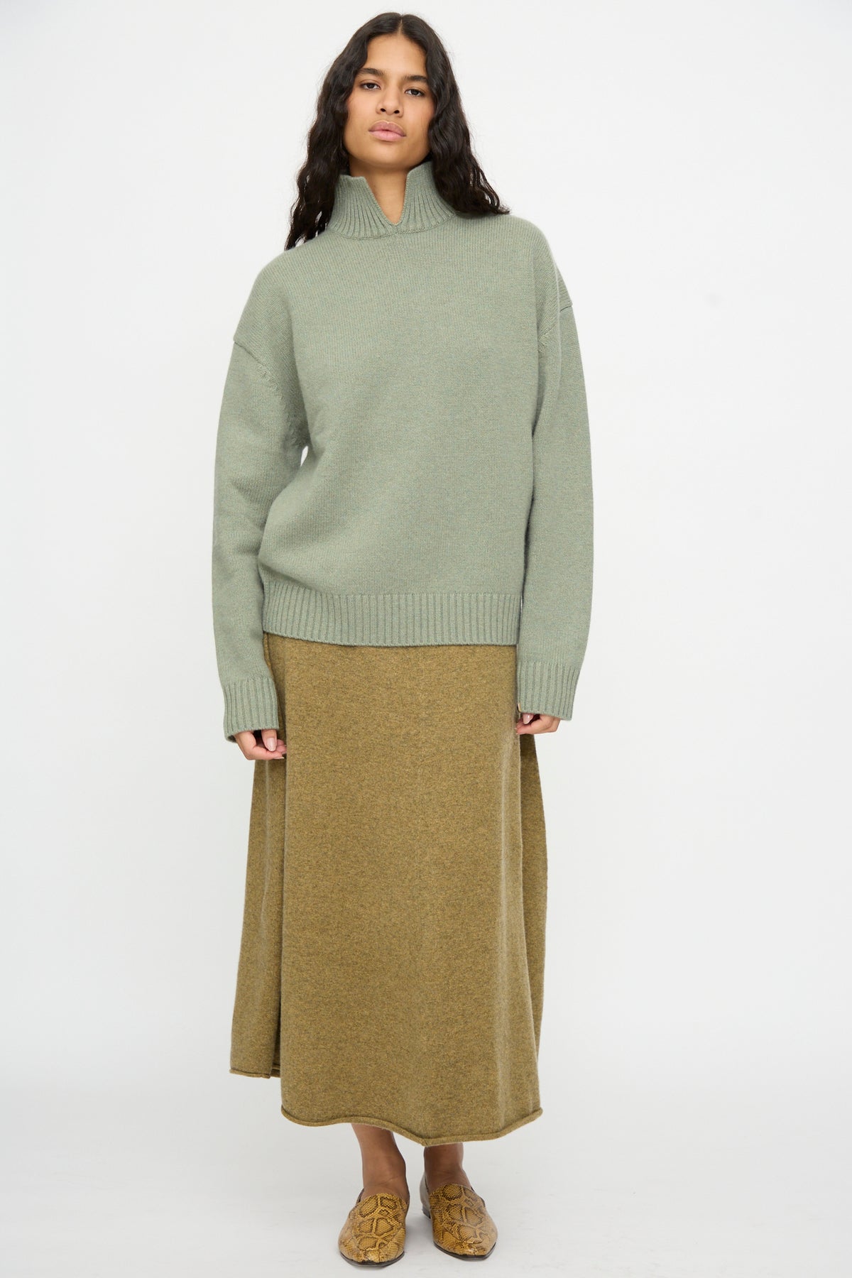 A woman stands facing forward against a plain white background, wearing the Extreme Cashmere No. 514 Jan Sweater in Hemlock, paired with an olive maxi skirt and yellow patterned slip-on shoes.