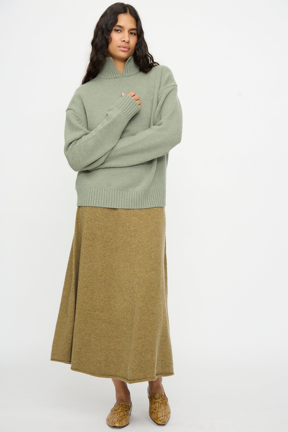 A person stands against a white background wearing the Extreme Cashmere No. 514 Jan Sweater in Hemlock, paired with a tan ankle-length skirt and tan slip-on shoes.
