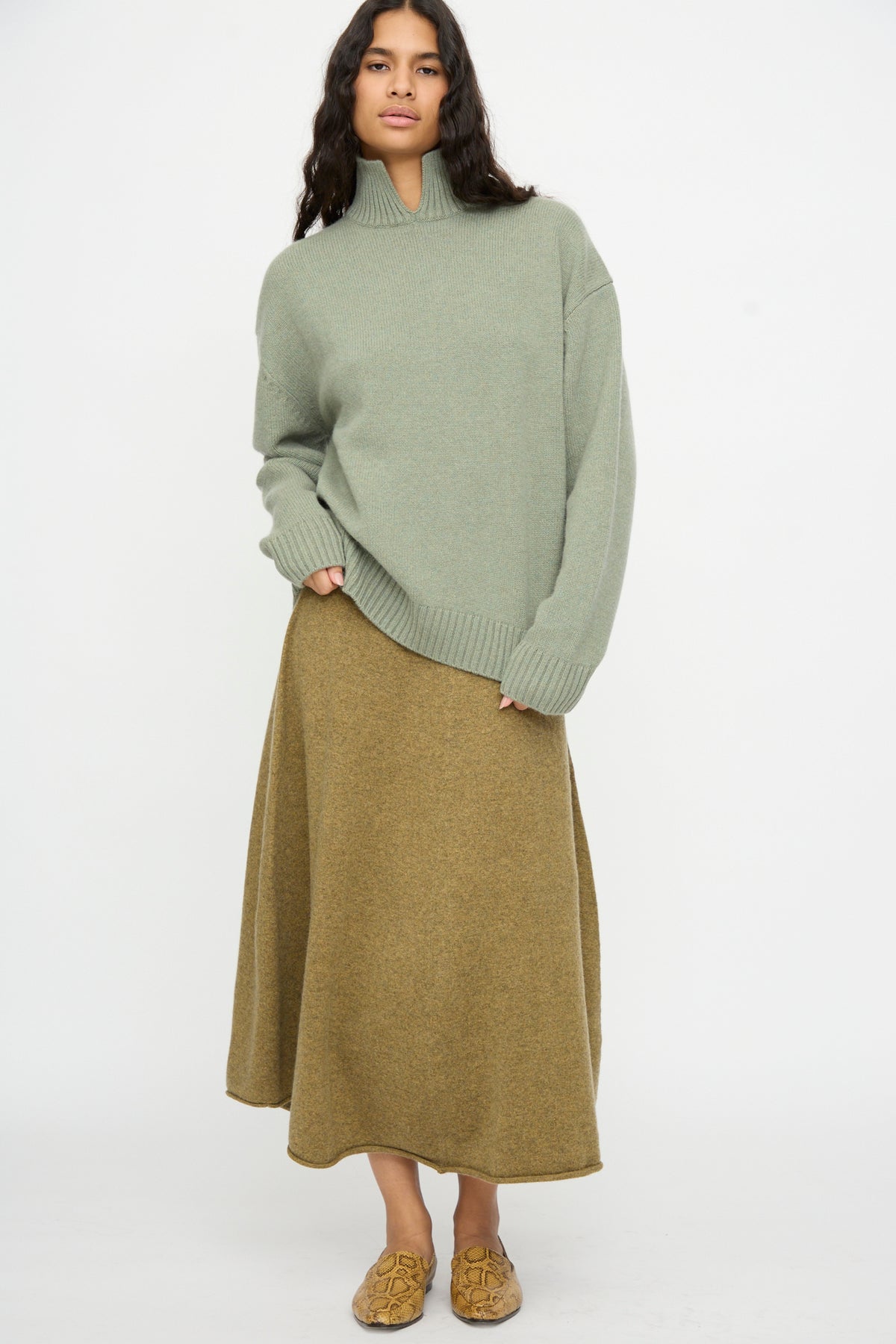 A person stands against a white background, wearing the No. 514 Jan Sweater in Hemlock by Extreme Cashmere, paired with an olive green skirt and tan loafers.