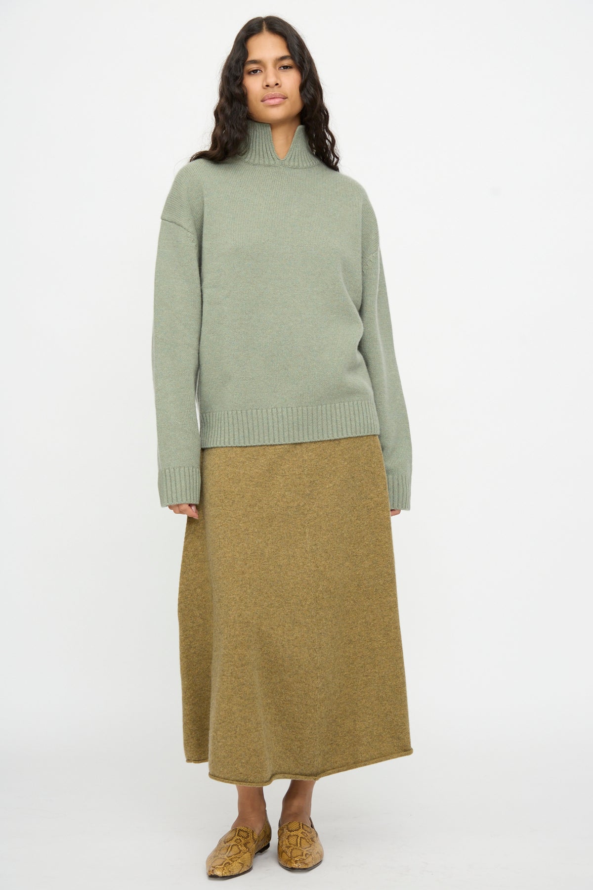 A person stands against a plain white background wearing the Extreme Cashmere No. 514 Jan Sweater in Hemlock, a brown maxi skirt, and tan patterned slip-on shoes.