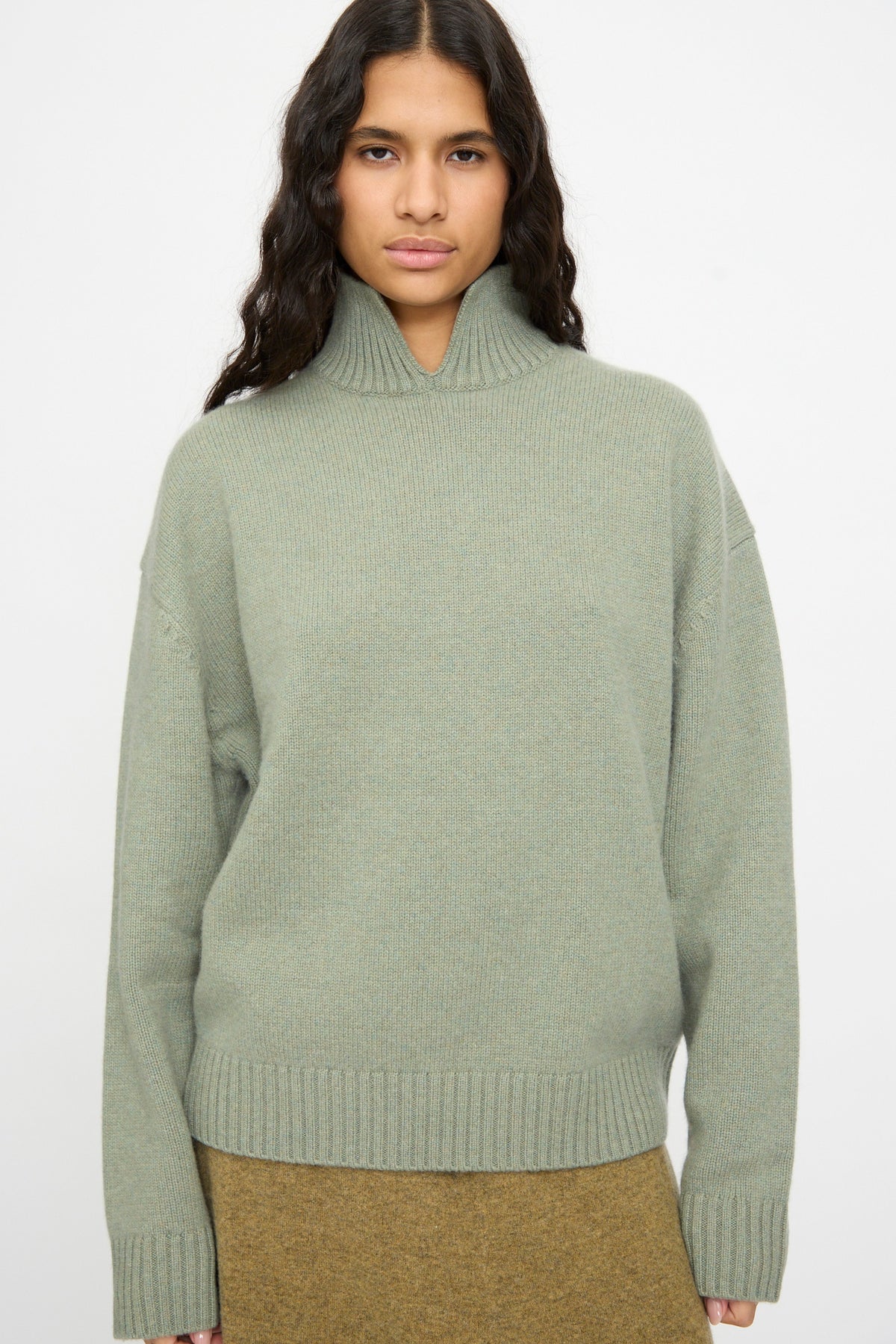 A person wearing the Extreme Cashmere No. 514 Jan Sweater in Hemlock, styled with an olive-brown skirt and standing against a plain white background.