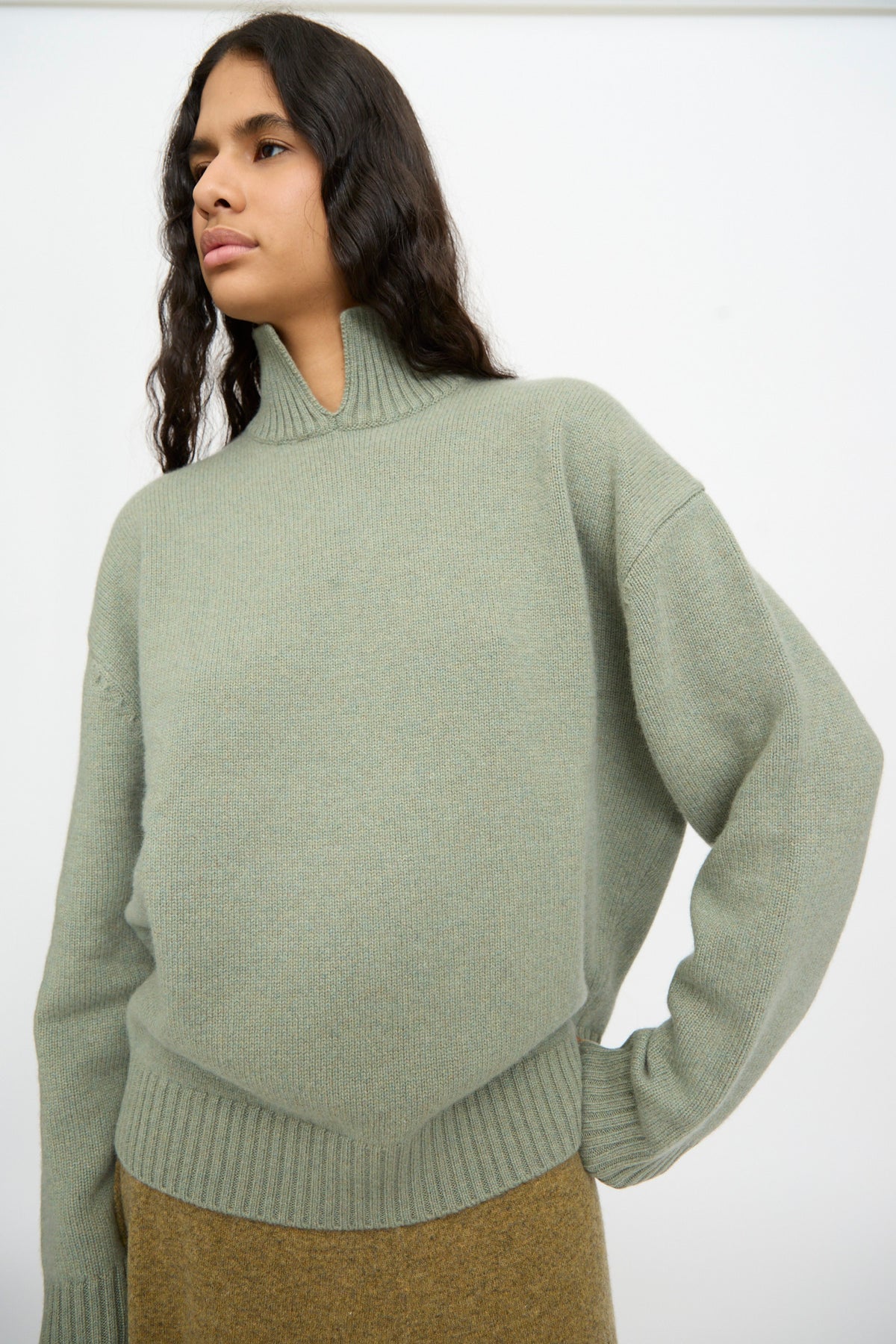 A person wearing the Extreme Cashmere No. 514 Jan Sweater in Hemlock, featuring ribbed cuffs and hem, stands in front of a plain white background.