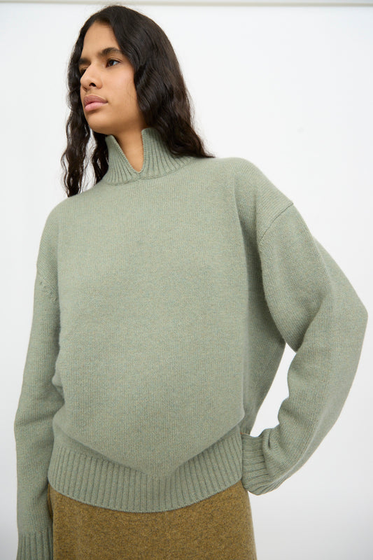 A person wearing the Extreme Cashmere No. 514 Jan Sweater in Hemlock, featuring ribbed cuffs and hem, stands in front of a plain white background.