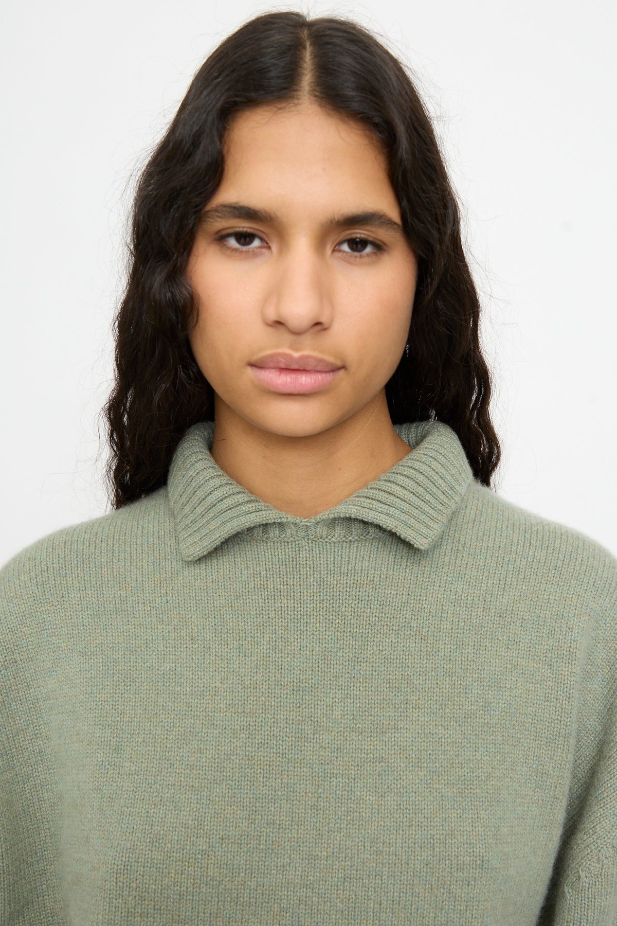 A person with long, wavy dark hair wears the Extreme Cashmere No. 514 Jan Sweater in Hemlock—a sage green cashmere sweater with a collar—and looks directly at the camera against a plain background.