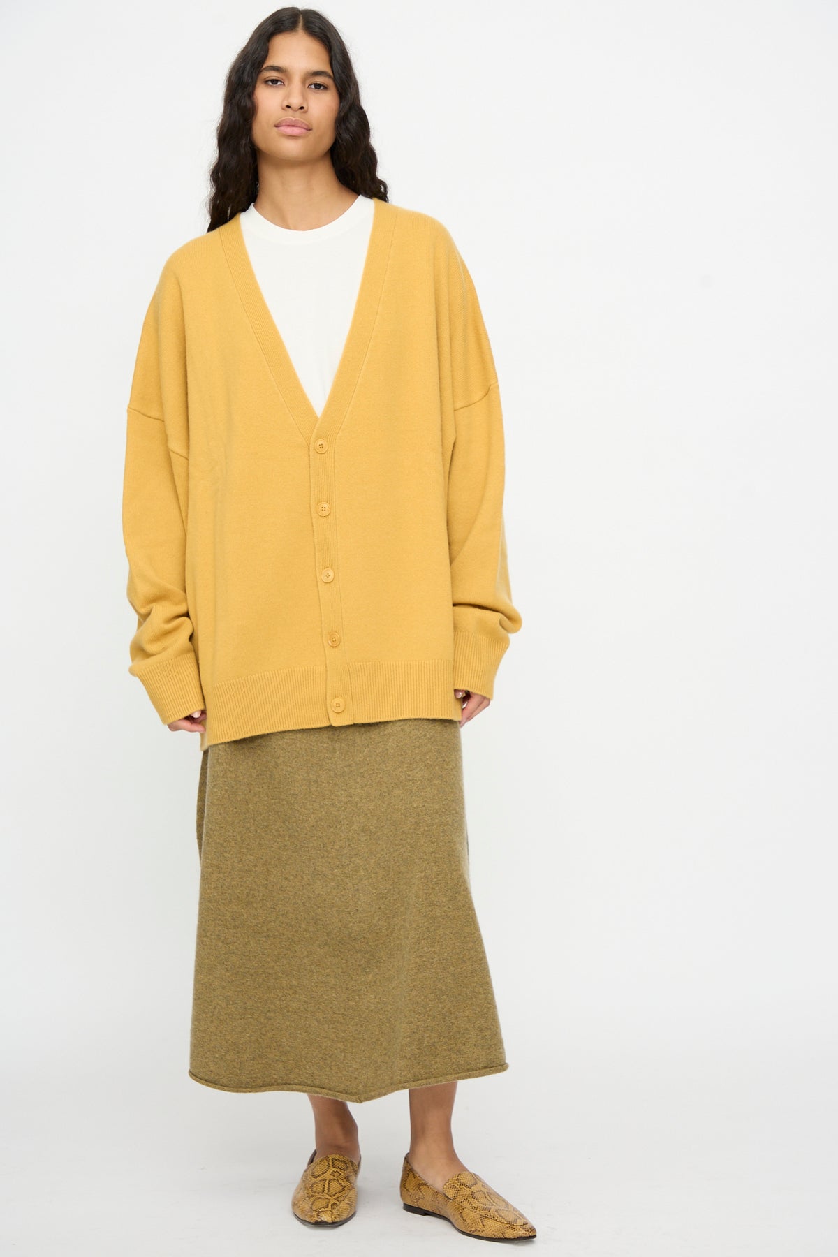 A person wearing the Extreme Cashmere No. 517 Simo Cardigan in Wheat—with baggy sleeves—layered over a white shirt, paired with an olive-colored long skirt and patterned shoes, stands against a plain white background.