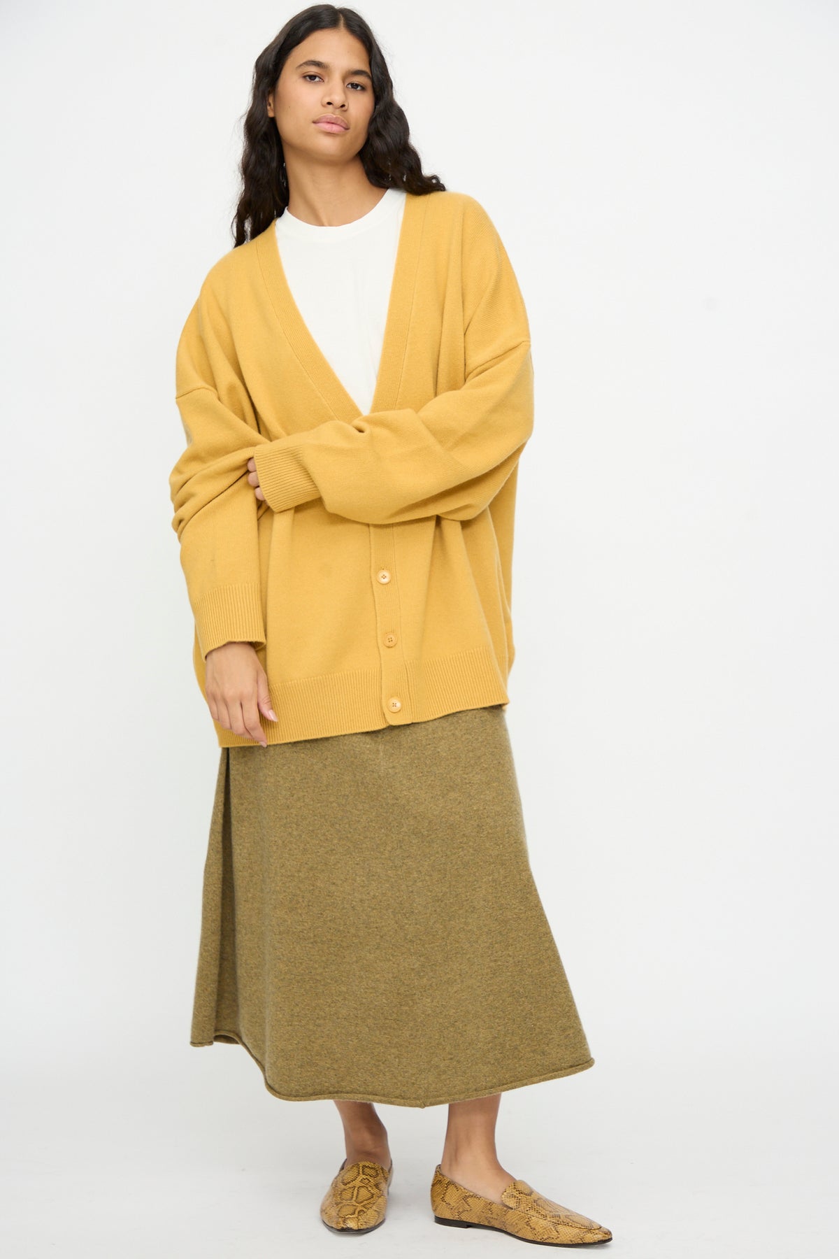 A person stands against a plain background wearing the Extreme Cashmere No. 517 Simo Cardigan in Wheat, a white shirt, an olive green skirt, and tan loafers.
