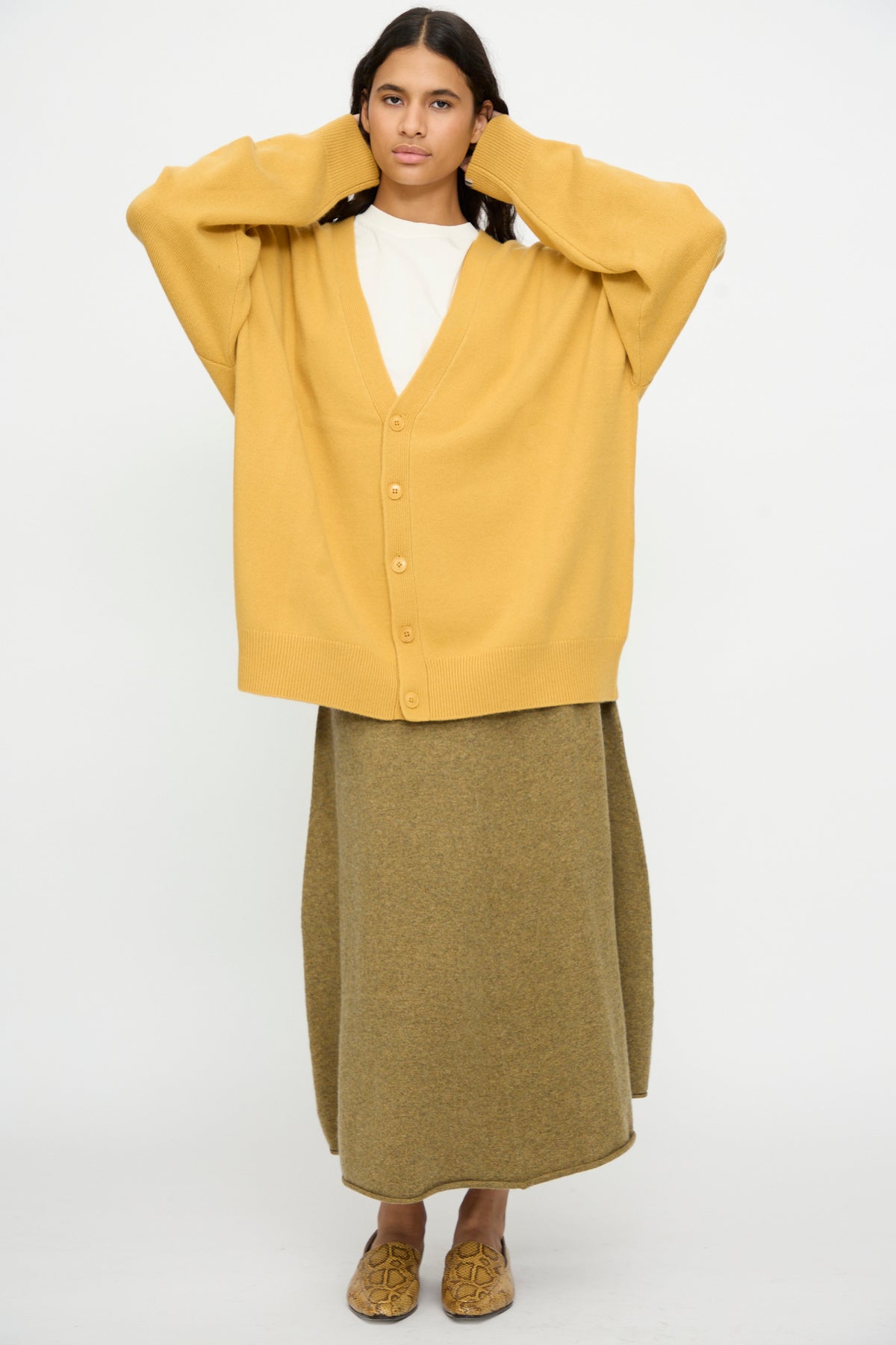 A person stands against a plain white background with hands touching head, wearing the Extreme Cashmere No. 517 Simo Cardigan in Wheat over a white top, paired with an olive green long skirt and tan loafers.