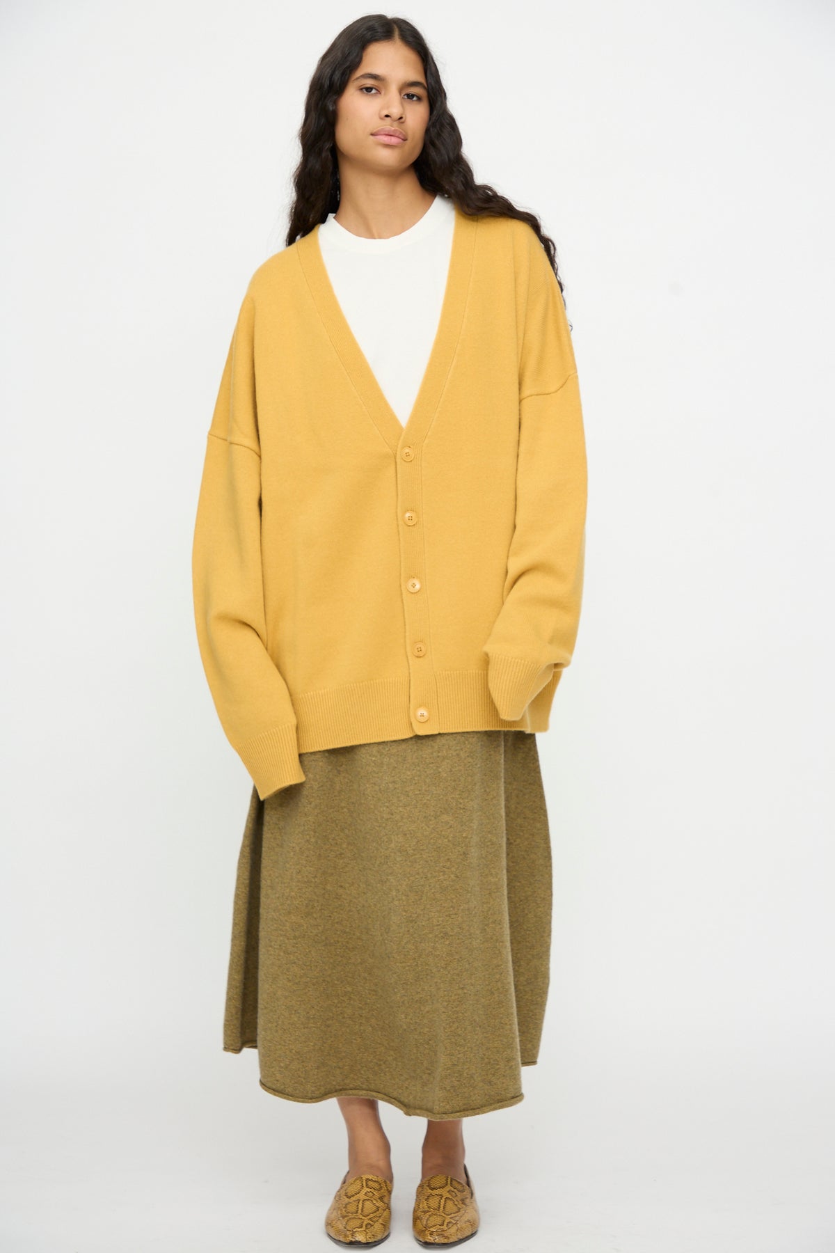 A woman stands against a plain background wearing the Extreme Cashmere No. 517 Simo Cardigan in Wheat, paired with a white top, an olive midi skirt, and patterned loafers.