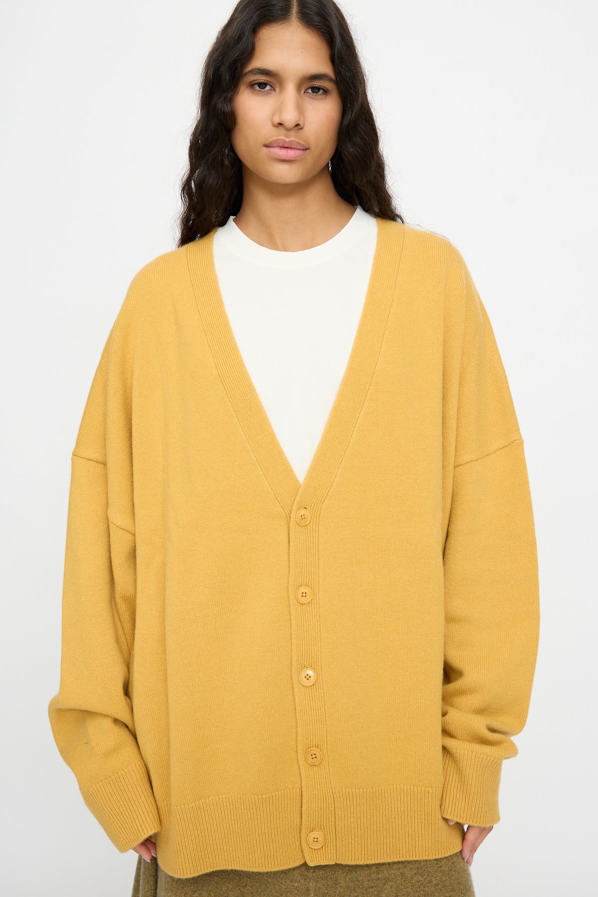 A person wearing the Extreme Cashmere No. 517 Simo Cardigan in Wheat, featuring an oversized fit and baggy sleeves, layered over a white shirt and standing against a plain light background.