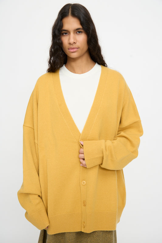 A person wearing the Extreme Cashmere No. 517 Simo Cardigan in Wheat—a loose-fitting women’s v-neck style—layered over a white shirt, stands against a plain light background.