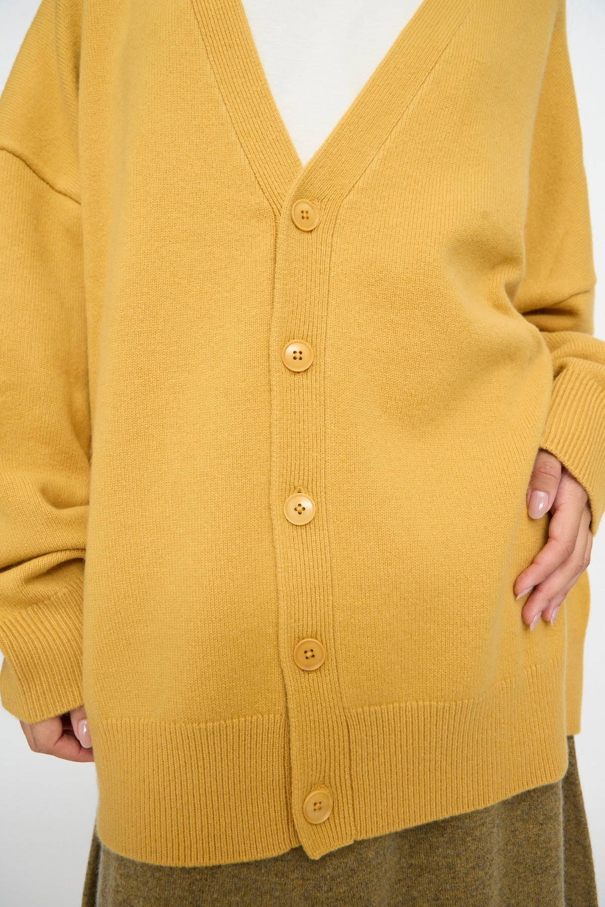 A person wearing the Extreme Cashmere No. 517 Simo Cardigan in Wheat, styled over a white top and brown skirt, stands against a plain background.