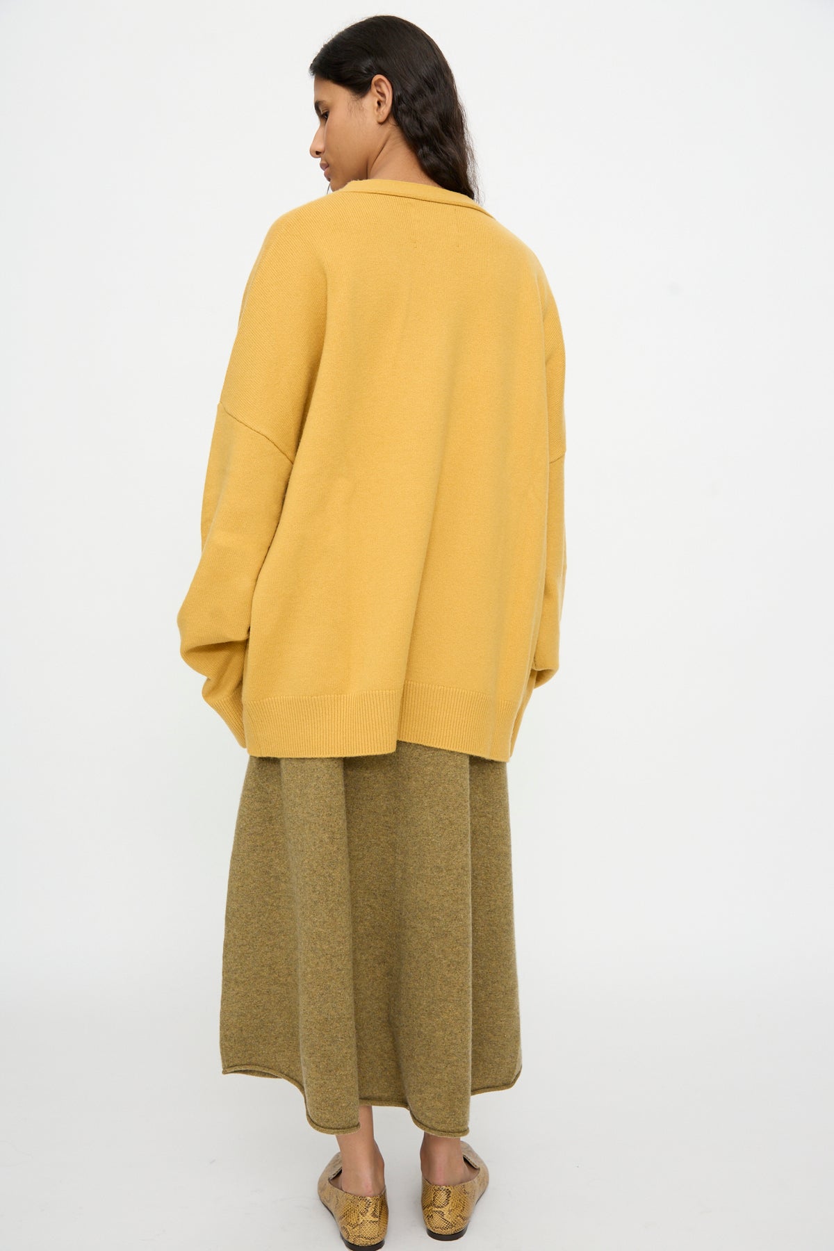 A woman stands with her back to the camera against a plain white background, wearing the Extreme Cashmere No. 517 Simo Cardigan in Wheat, paired with a long olive skirt and flat shoes.