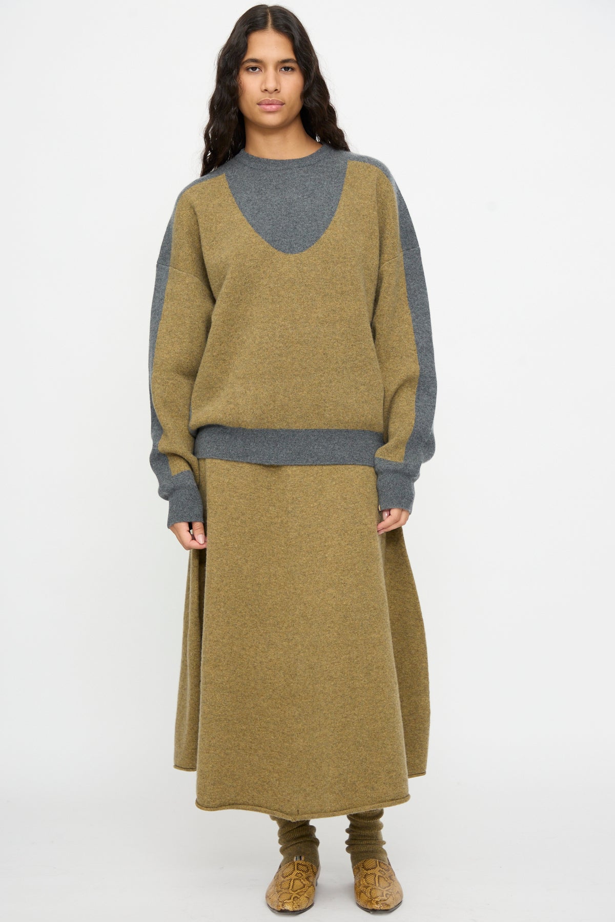 A person wearing the Extreme Cashmere No. 518 Trompe Sweater in Hunter and Felt—an oversized brown and gray knit with a crew neckline—paired with a matching brown skirt, socks, and shoes, standing against a plain white background.