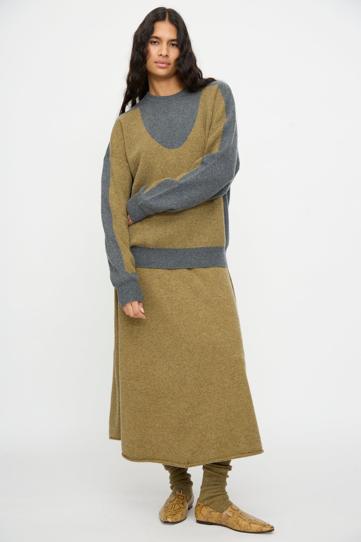 Standing against a plain white background, the person is wearing the Extreme Cashmere No. 518 Trompe Sweater in Hunter and Felt—a loose-fitting knit sweater with a crew neckline—paired with a matching long skirt in coordinating gray and olive tones, matching leggings, and beige shoes.