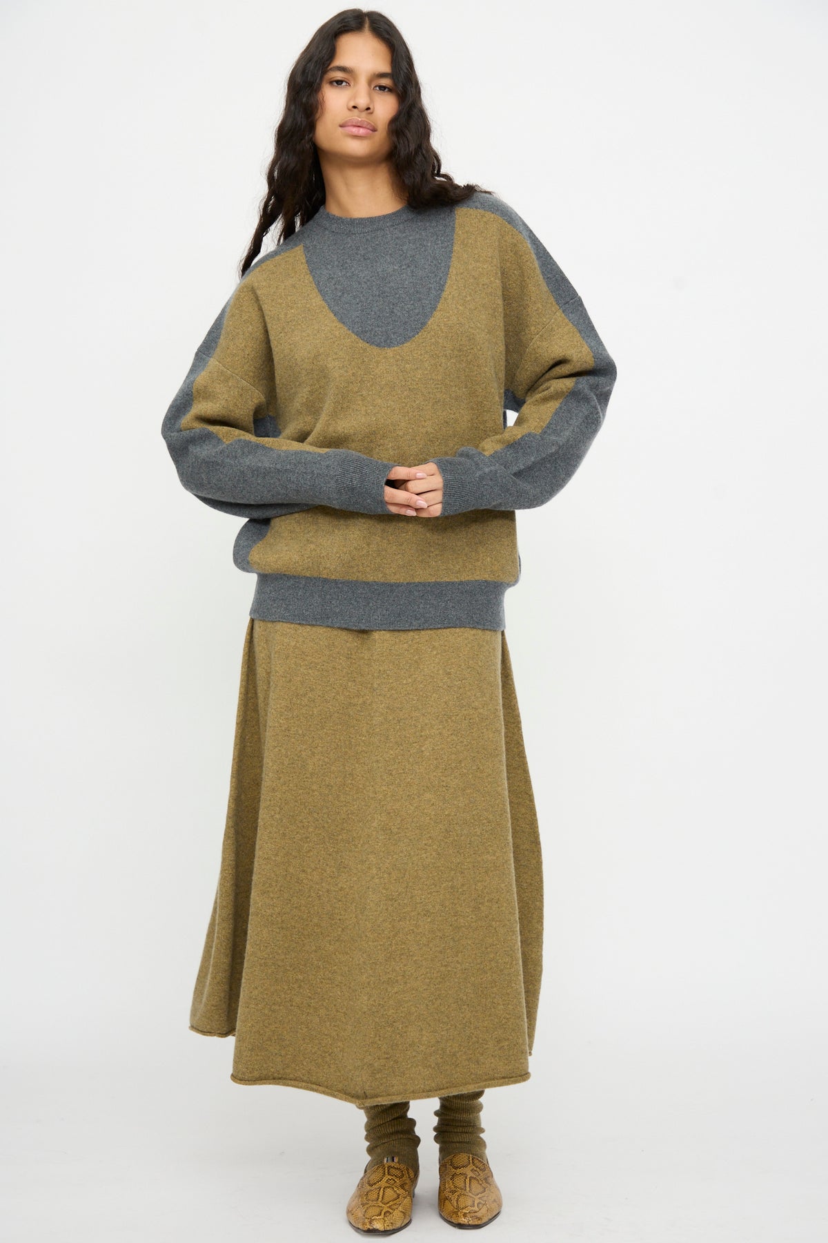 A person wearing the Extreme Cashmere No. 518 Trompe Sweater in Hunter and Felt—a loose-fitting, colorblock knit with a crew neckline—paired with a matching long skirt and patterned socks, stands against a plain white background.